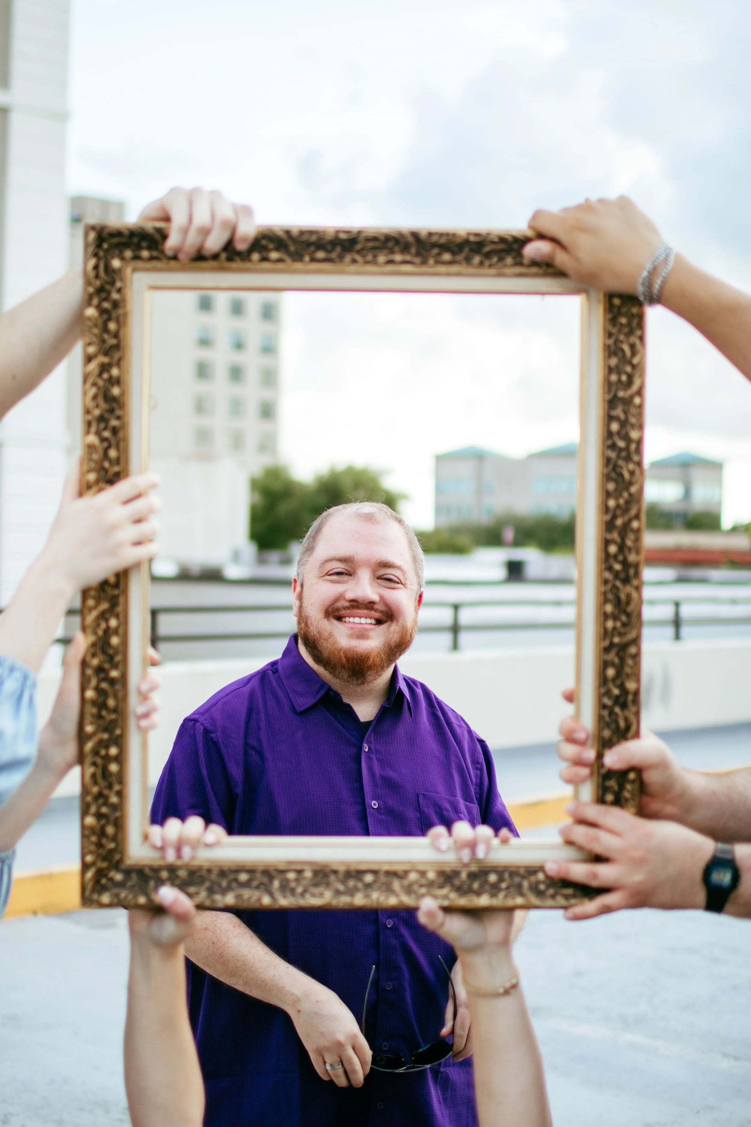 A man with a beard and a purple shirt smiling while looking through a decorative frame held by several people outdoors on a cloudy day.