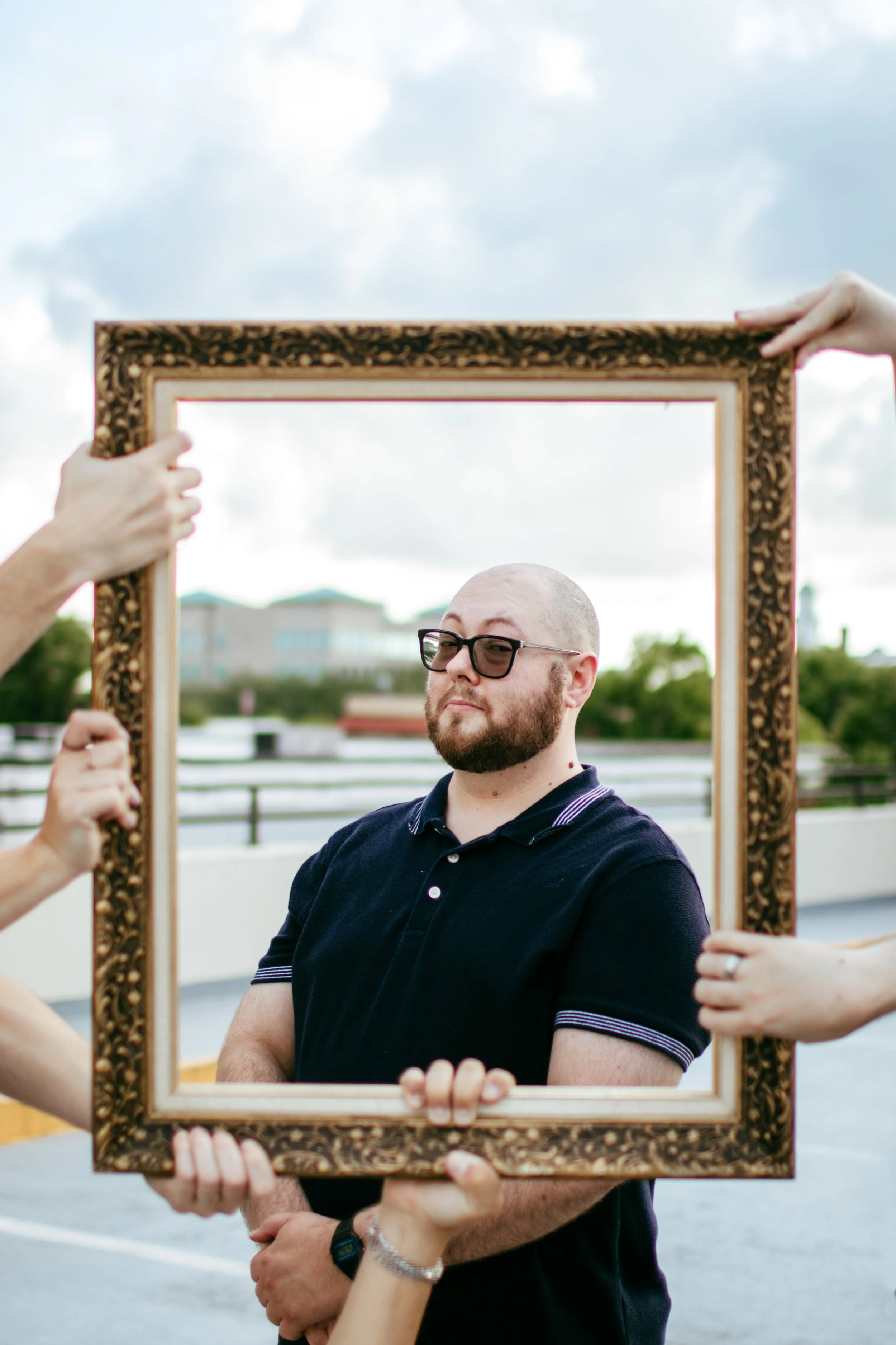 A man with glasses and a beard posing behind an ornate picture frame being held by four people outside on a cloudy day.