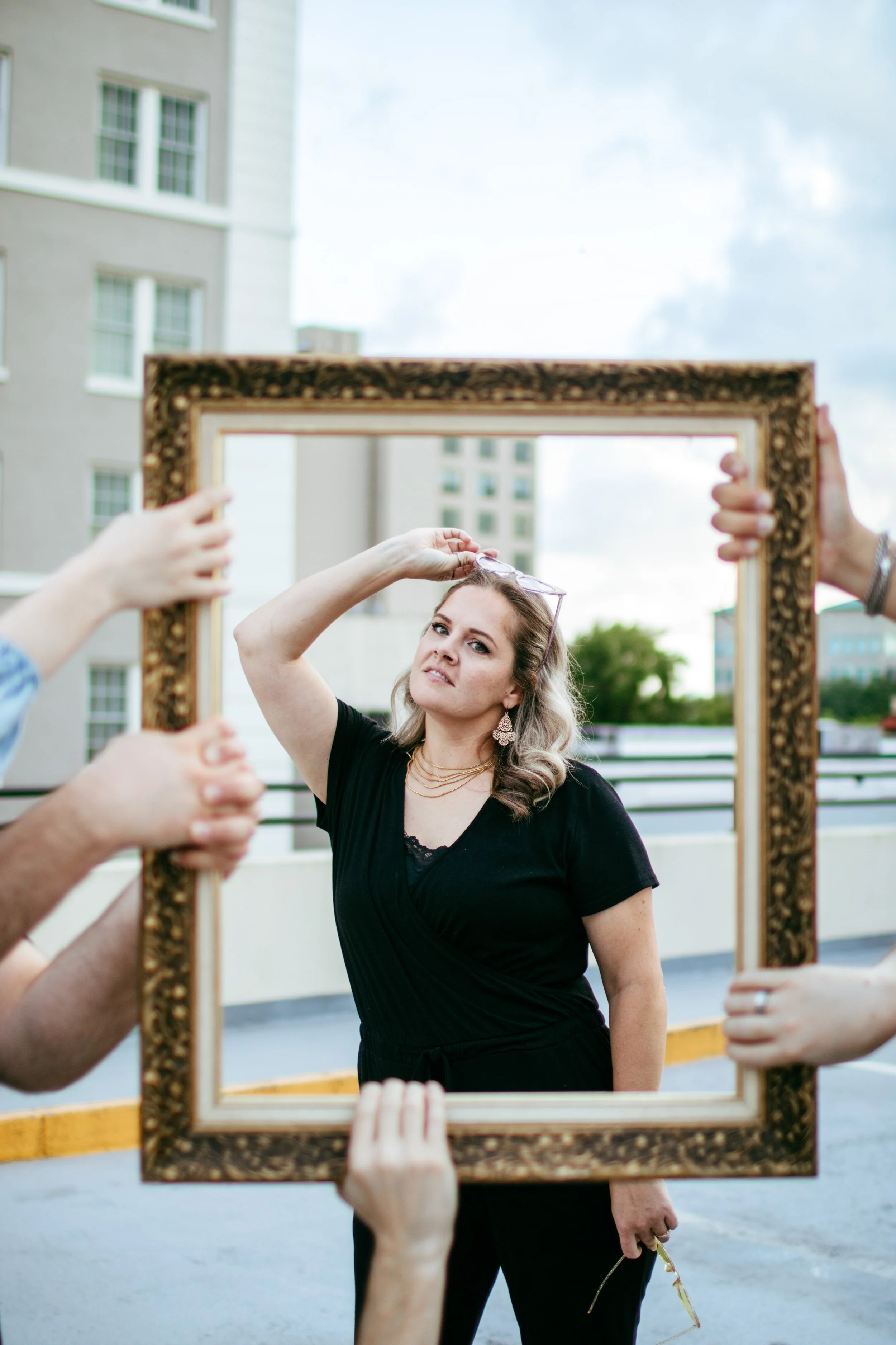 Woman in black dress posing with sunglasses on her head while holding a pair of glasses in her right hand during photo shoot on rooftop, framed by a decorative picture frame held by multiple hands.