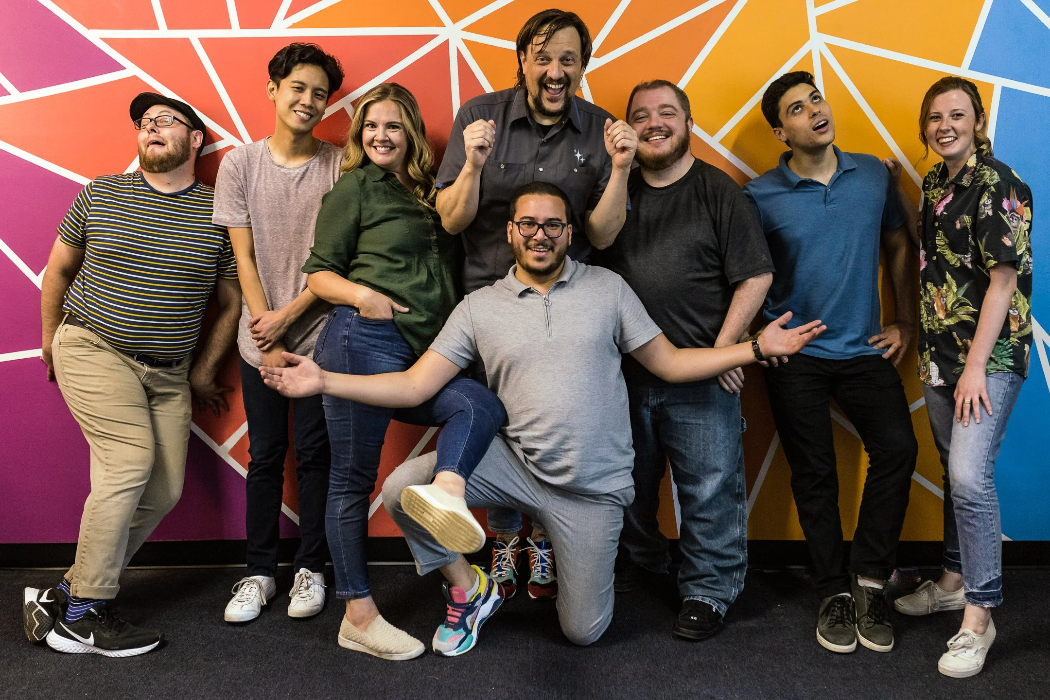 A diverse group of nine people posing together against a colorful geometric mural wall. They are smiling and making playful gestures, dressed casually.