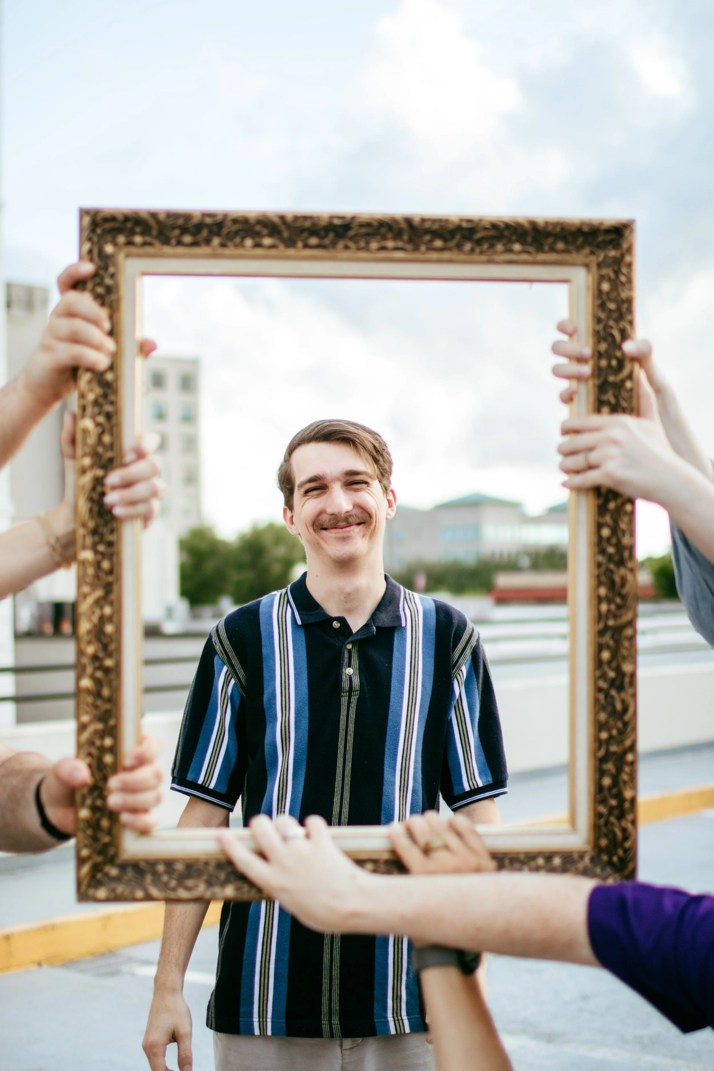 A smiling young man with a mustache stands outdoors, seen through a decorative wooden photo frame held by four people, with buildings and a cloudy sky in the background.