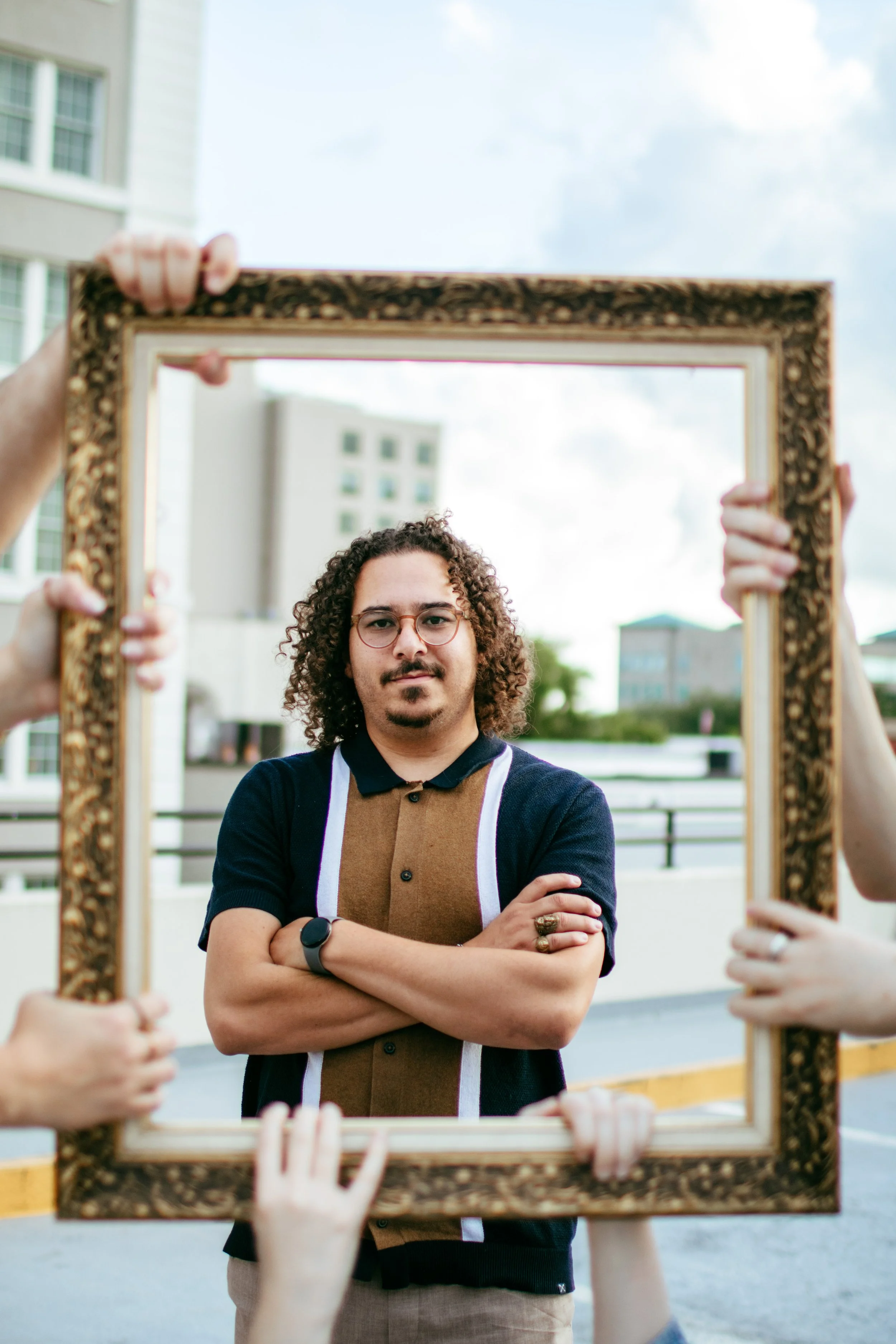 A man with curly hair, glasses, and a mustache standing outdoors with arms crossed, framed by several hands holding an ornate picture frame.