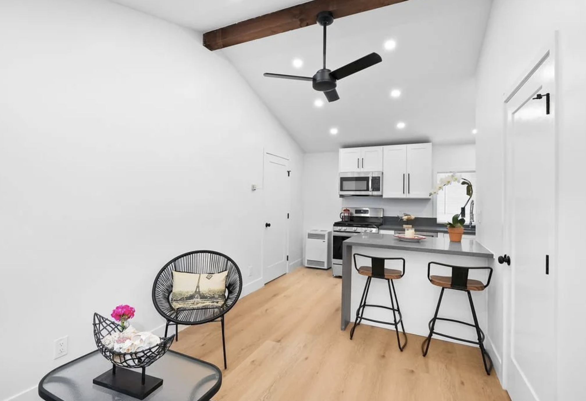Modern kitchen and dining area with white walls, ceiling fan, recessed lighting, black and wood chairs, small table with pink flowers, and kitchen appliances including microwave, oven, and a potted plant on the counter.