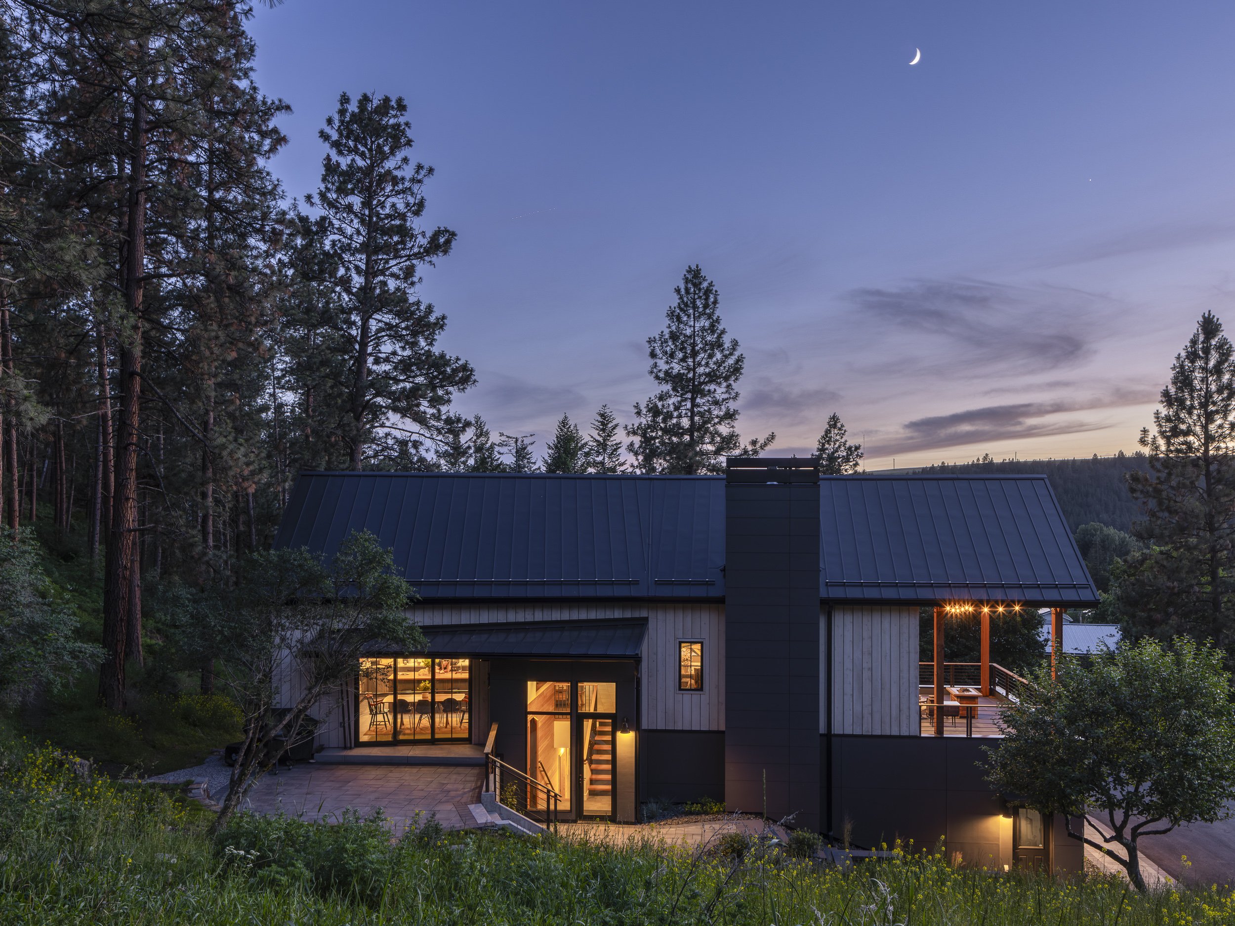 Modern house with large glass windows and a patio, surrounded by trees, during twilight with a visible crescent moon and stars.