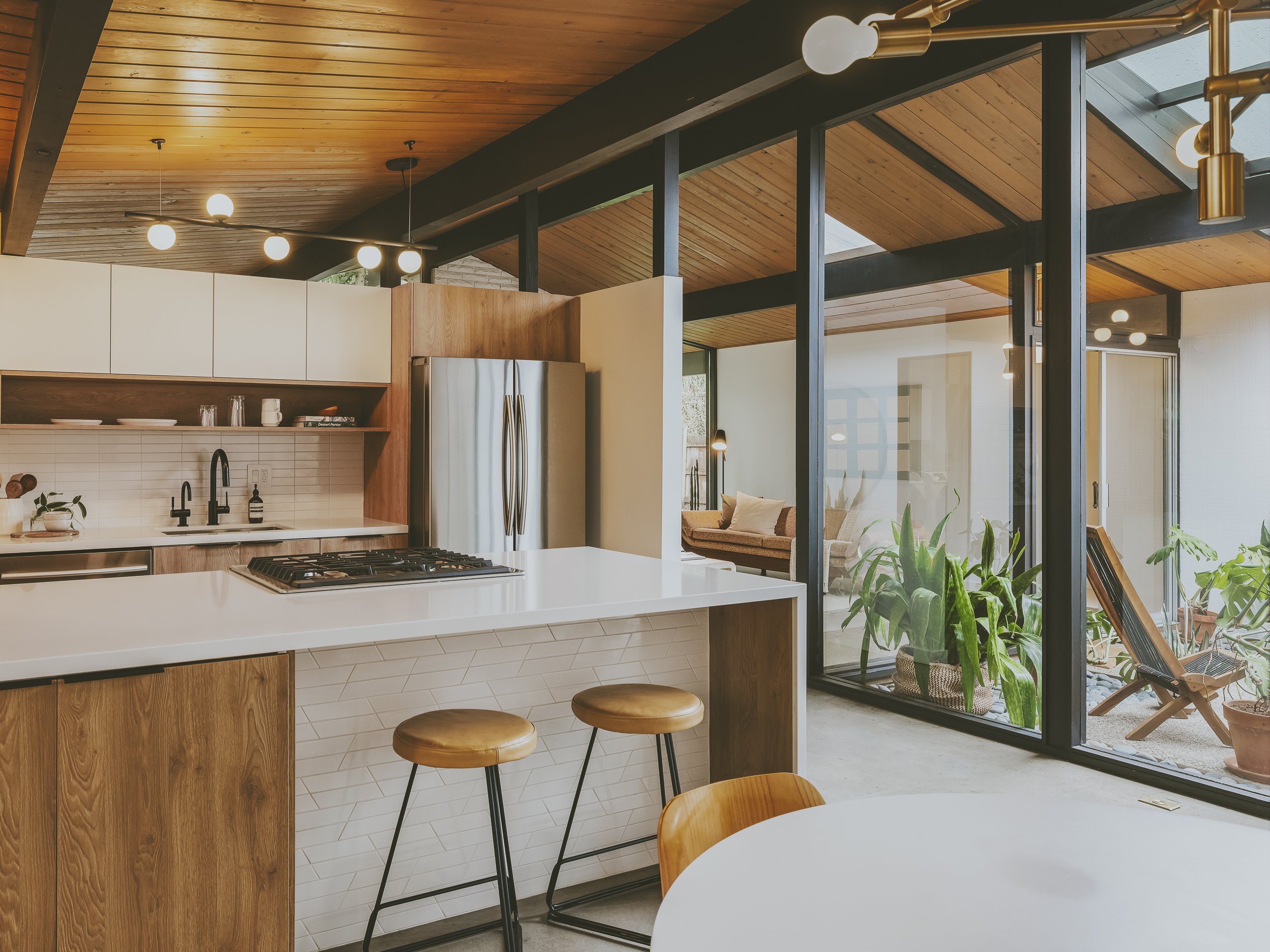 Modern kitchen with white countertops, wooden accents, and large glass sliding doors leading to a sunlit outdoor patio with plants and seating.