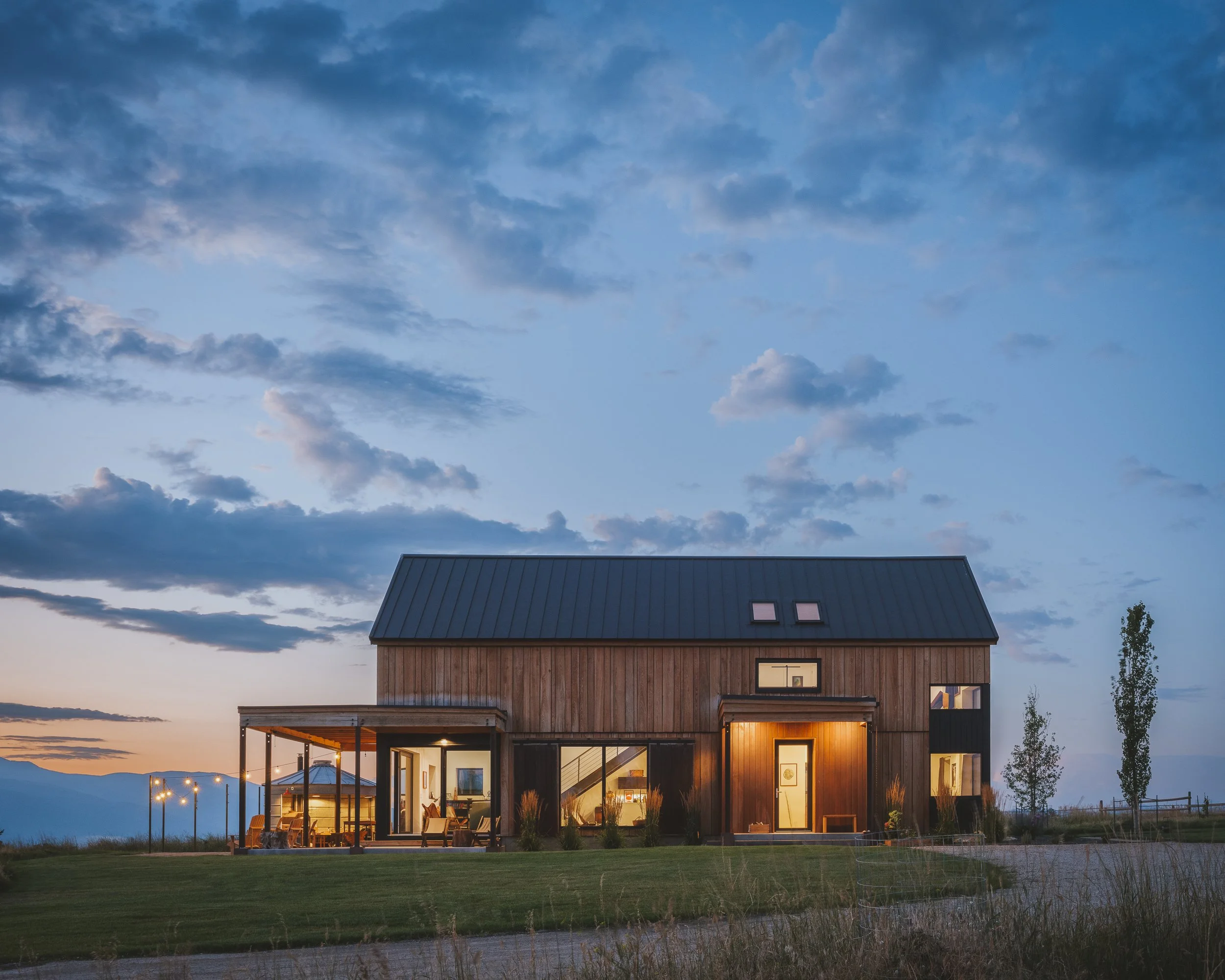 A modern two-story wooden house with large windows, illuminated from inside, under a partly cloudy sky at dusk. The house features a covered patio with outdoor seating and a lawn in the foreground.