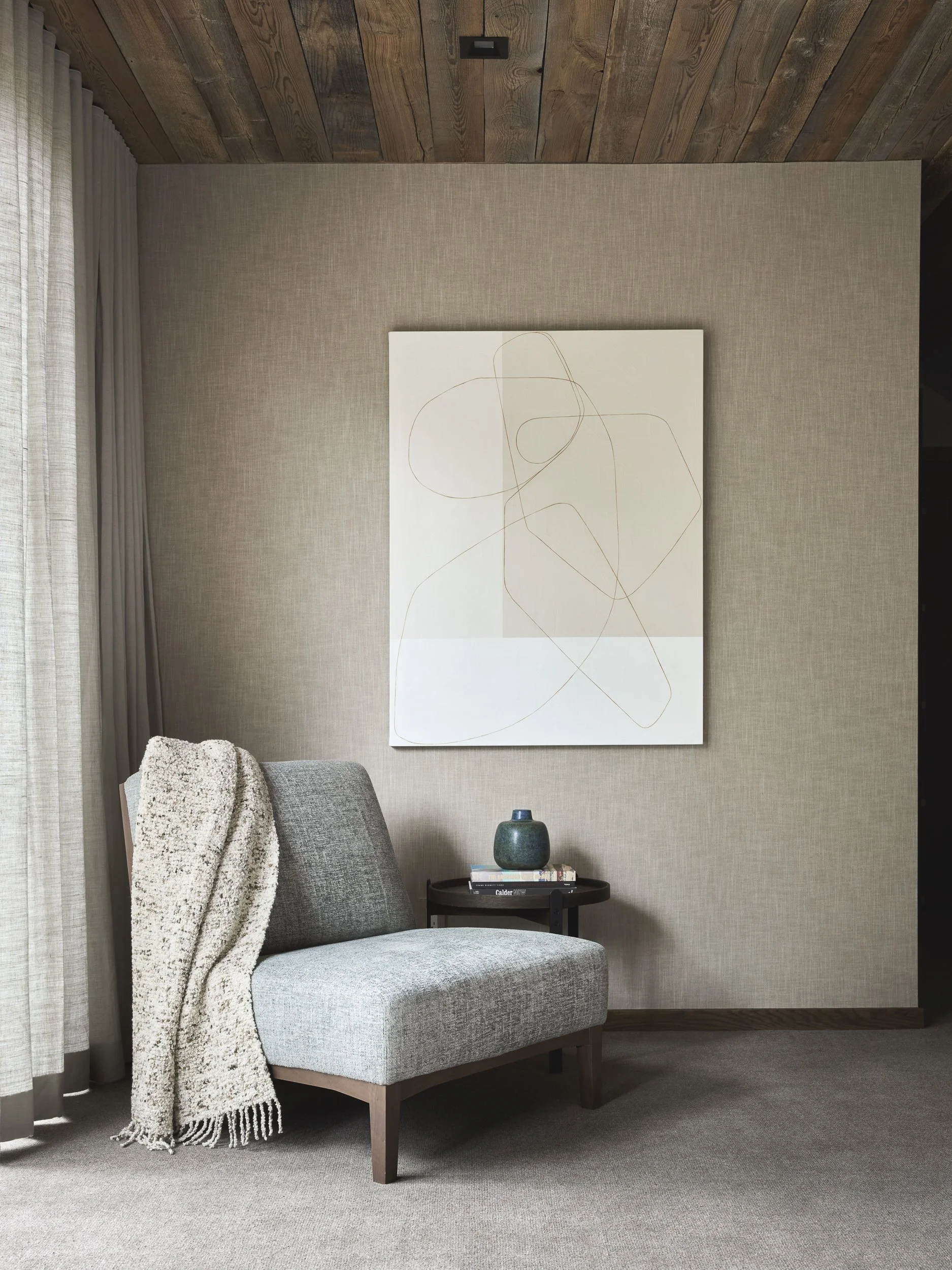 A cozy corner with a gray upholstered armchair draped with a cream throw blanket, a small black side table with a green vase and books, beige textured wall art, beige textured walls, and a wooden ceiling with sliding curtains.