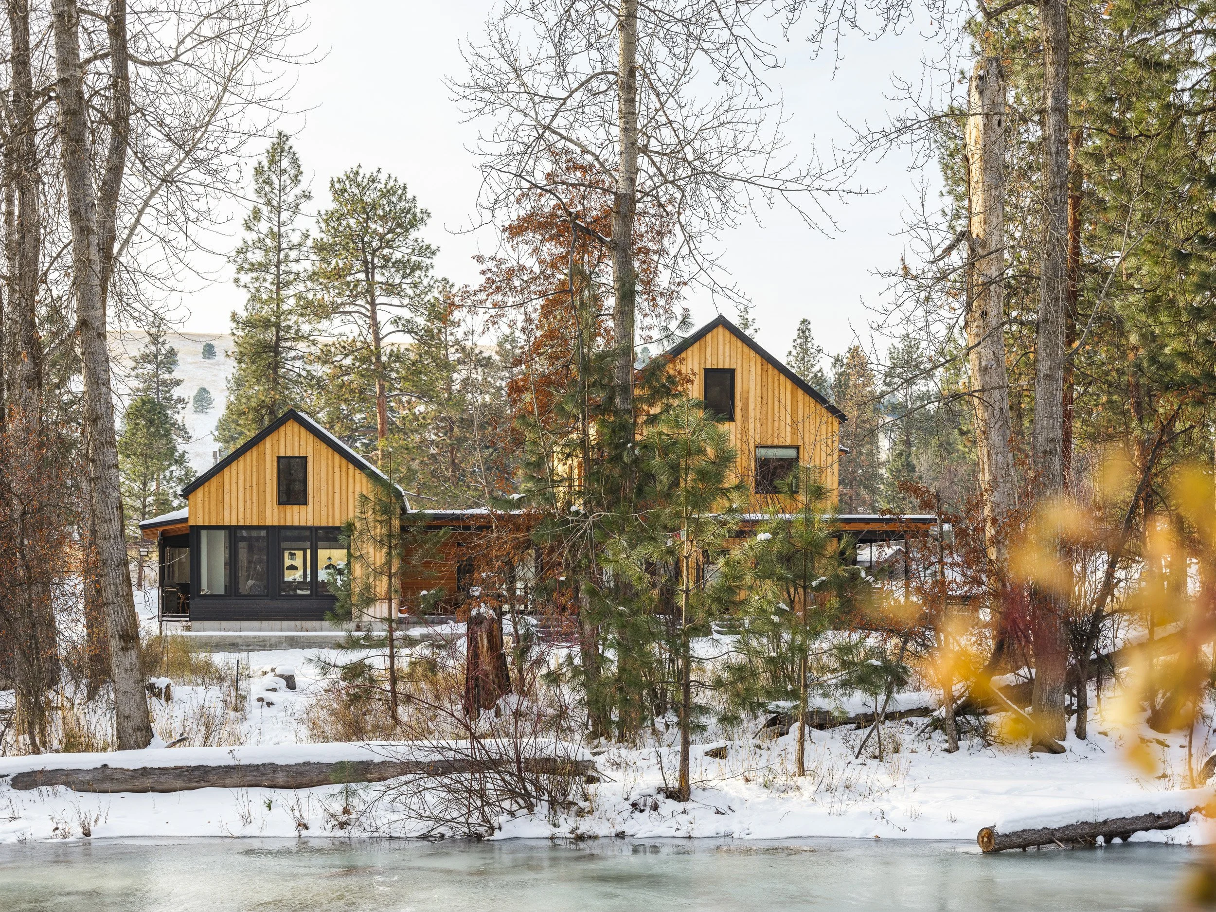 A modern house with wooden siding and large windows situated in a snowy forest next to a small body of water.