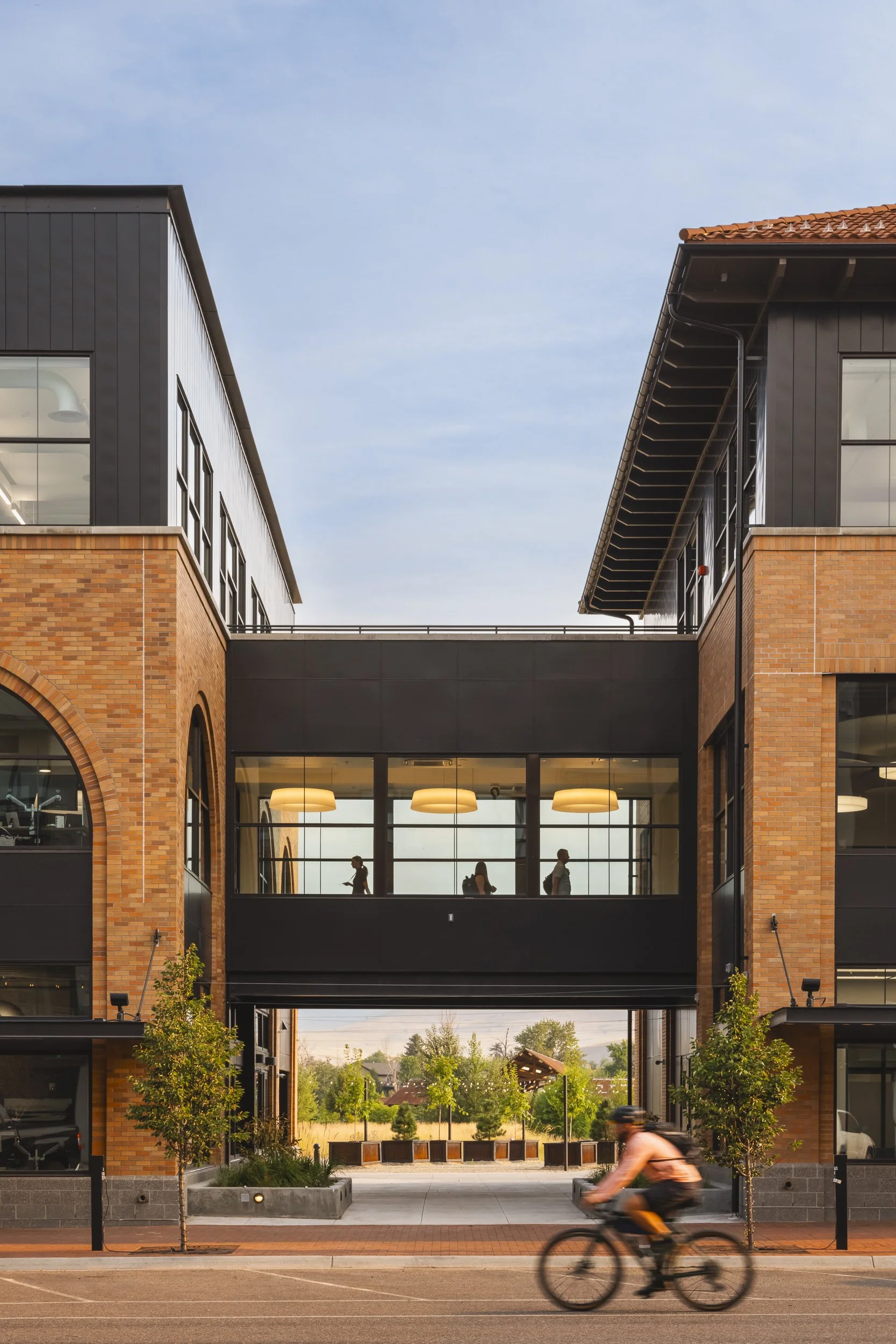 Modern building with brick and black siding, featuring large windows and a glass skybridge with silhouettes of people walking inside. A cyclist rides by on the street in front of the building, and trees line the sidewalk.