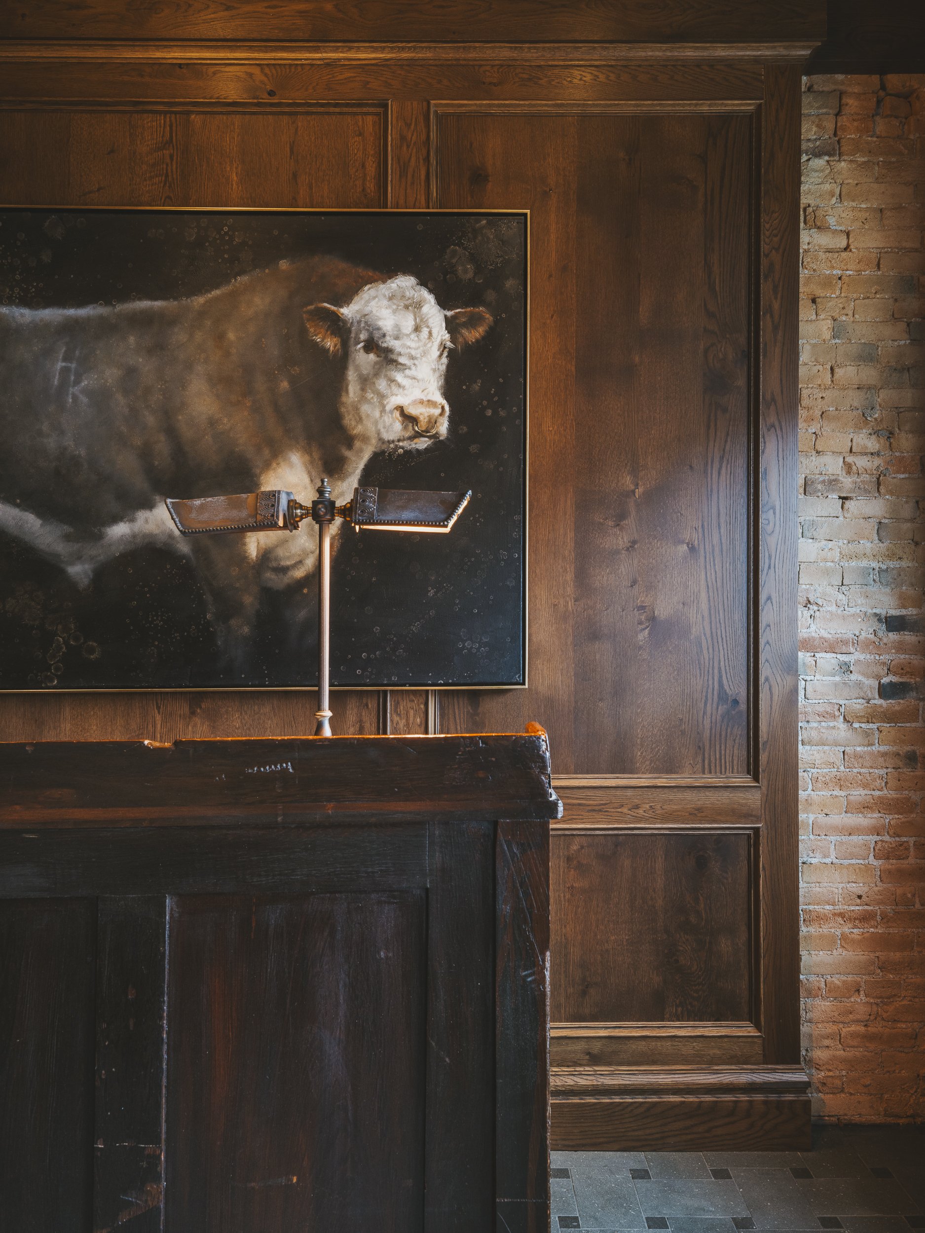 A painting of a cow on a dark wall behind a wooden counter in a room with brick and wood-paneled walls.