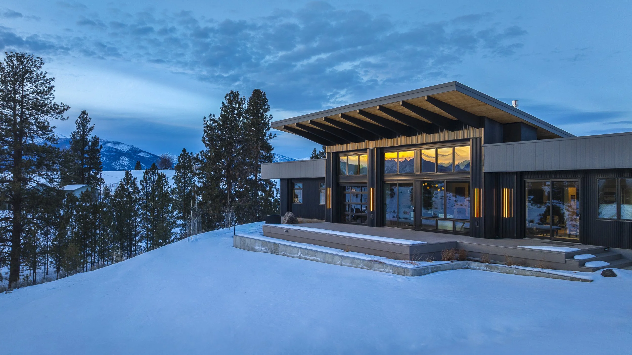Modern house with large glass windows and wooden accents in a snowy landscape at sunset, surrounded by pine trees and mountains.