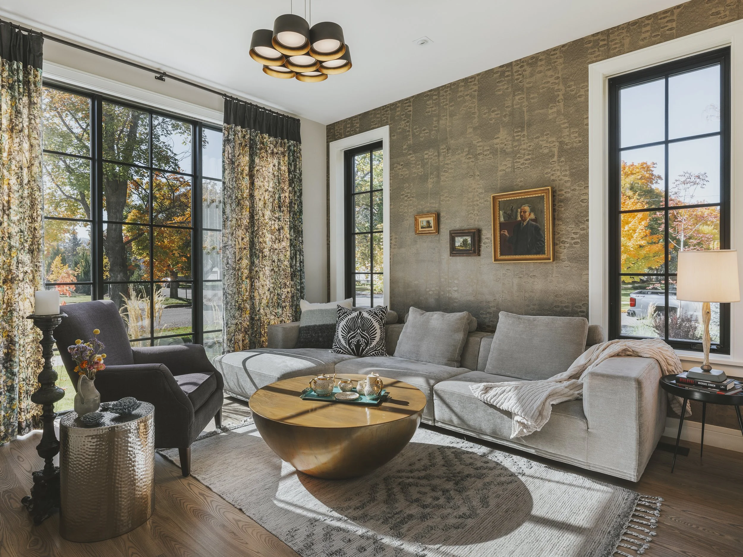 Living room with large window, decorated couch, armchair, coffee table with tea set, lamp, framed paintings, and curtains with autumn foliage views.