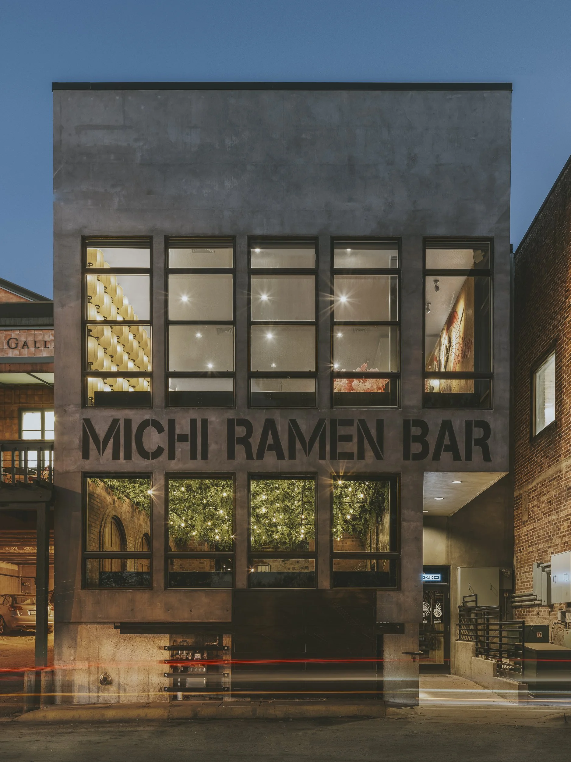 The exterior of a building at dusk with a sign reading 'MICH RAMEN BAR.' The building has large windows revealing the lit interior with ceiling lights and decor, and a small alleyway on the right side.