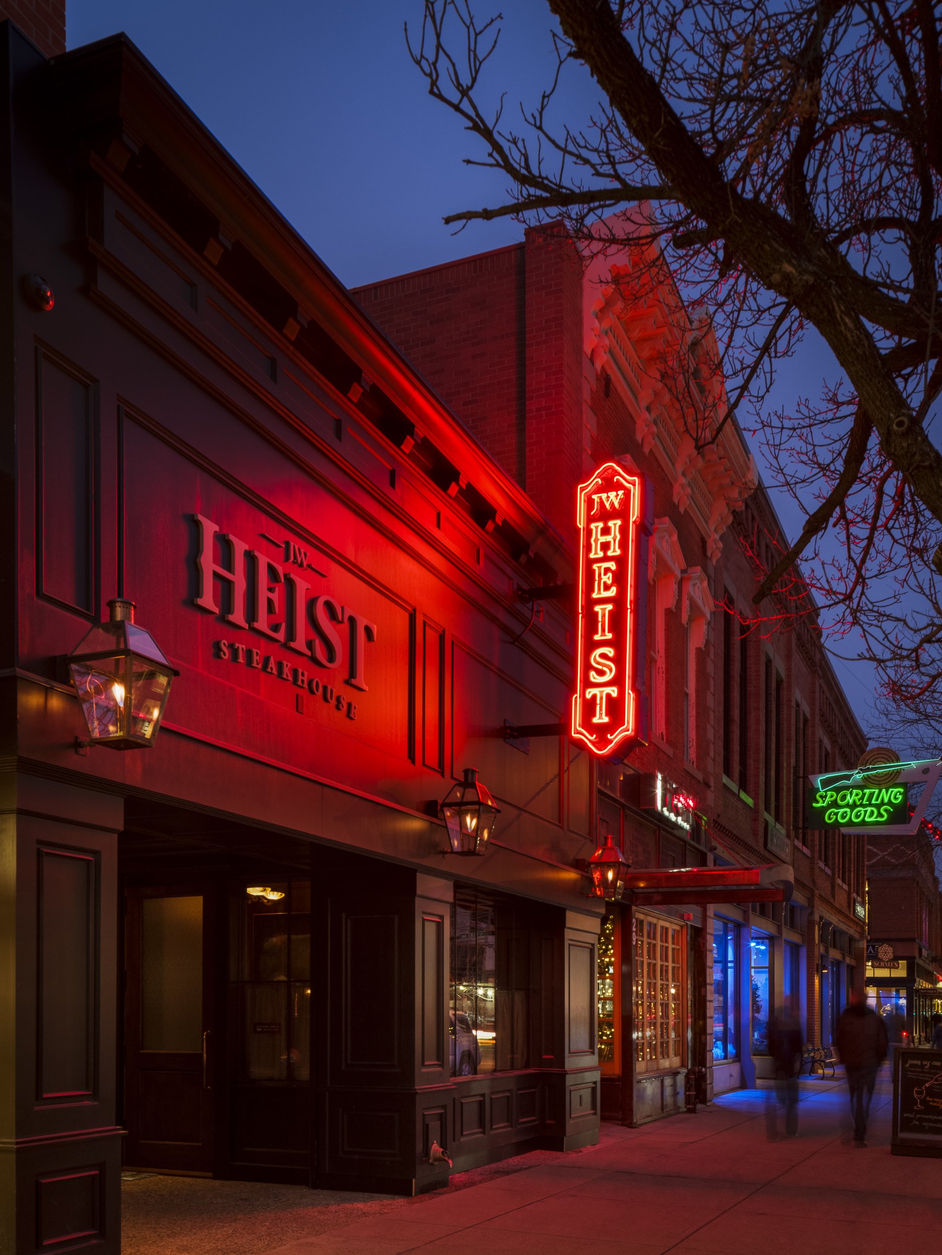 Street view of a restaurant called 'JW Heist Steaksehouse' with red neon signage, black exterior, and lantern-style lights, with people walking on the sidewalk and a leafless tree in the foreground.