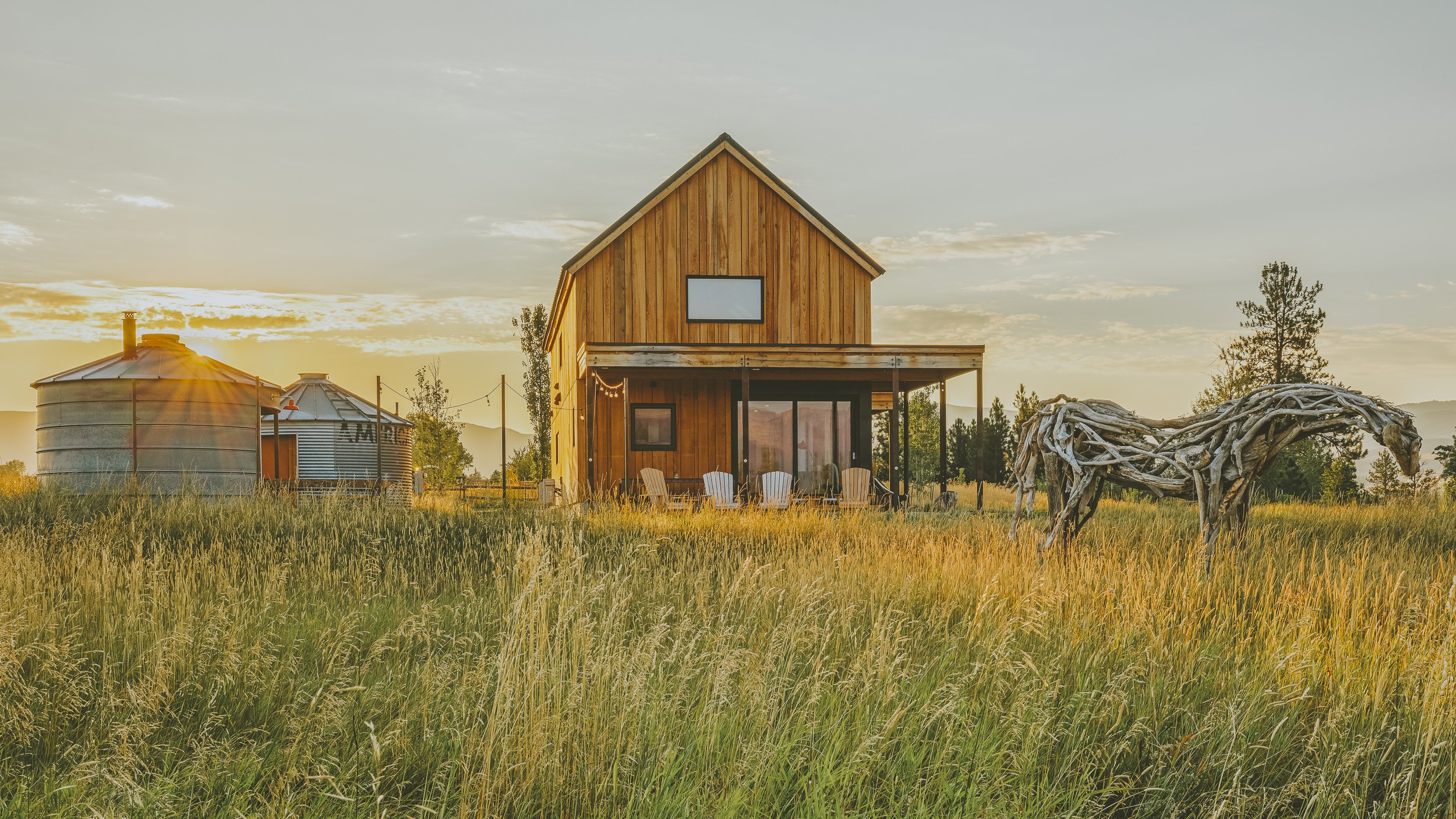 A wooden house with a porch and chairs, two metal grain silos, and a driftwood sculpture of a horse in a grassy field during sunset.