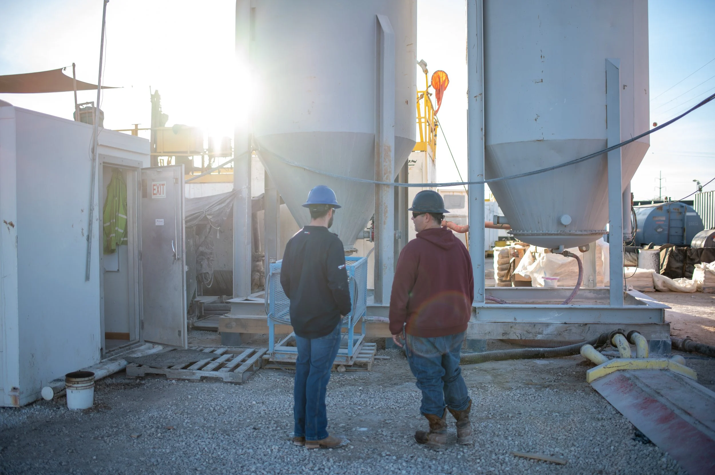 Two construction workers wearing helmets and work clothes are standing on a gravel surface at an industrial site, with large metal storage silos and equipment behind them. The sun is shining brightly, creating a glare in the background.