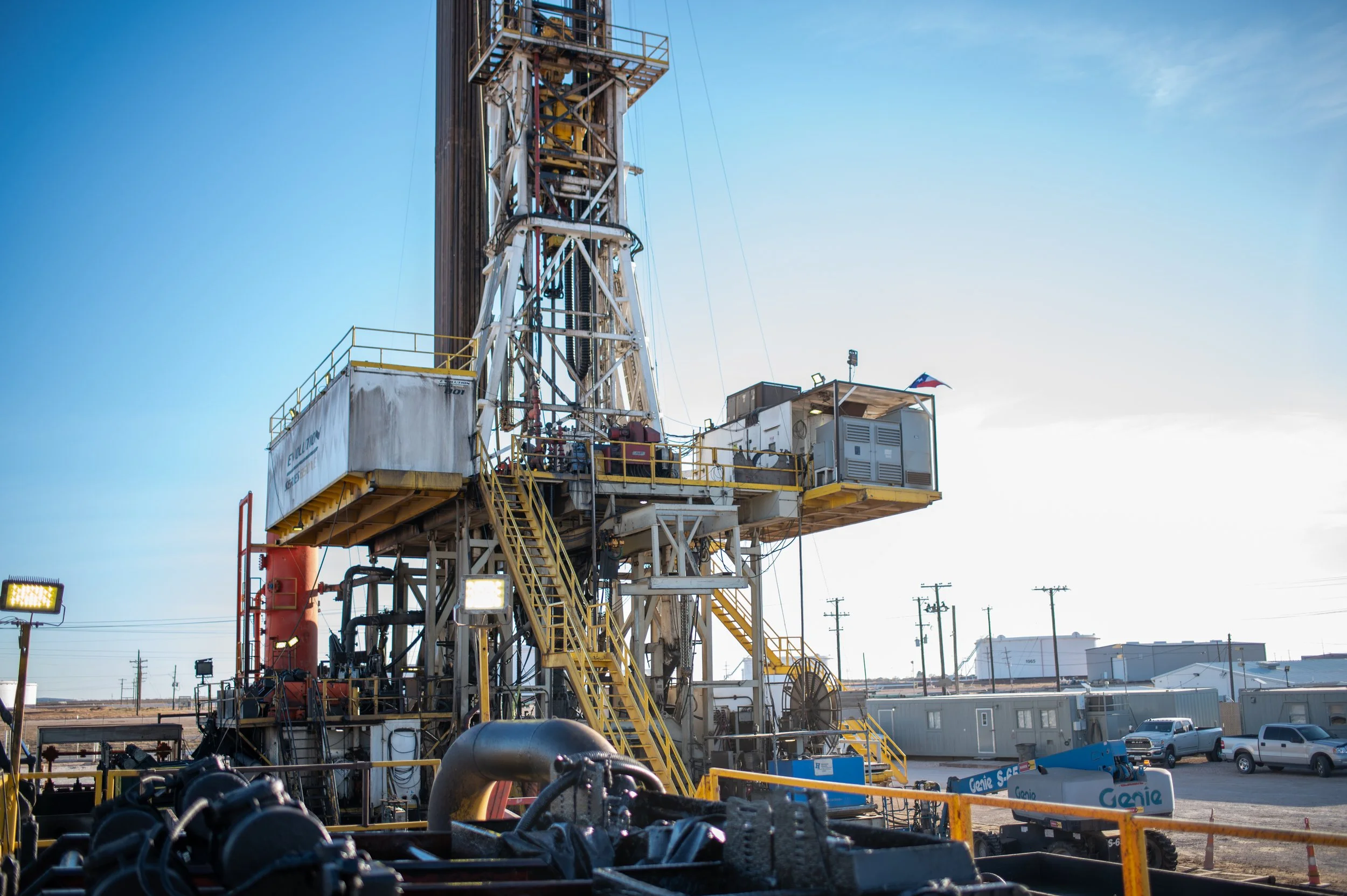 Oil drilling rig set up outdoors under a clear sky with parked cars and industrial buildings in the background.