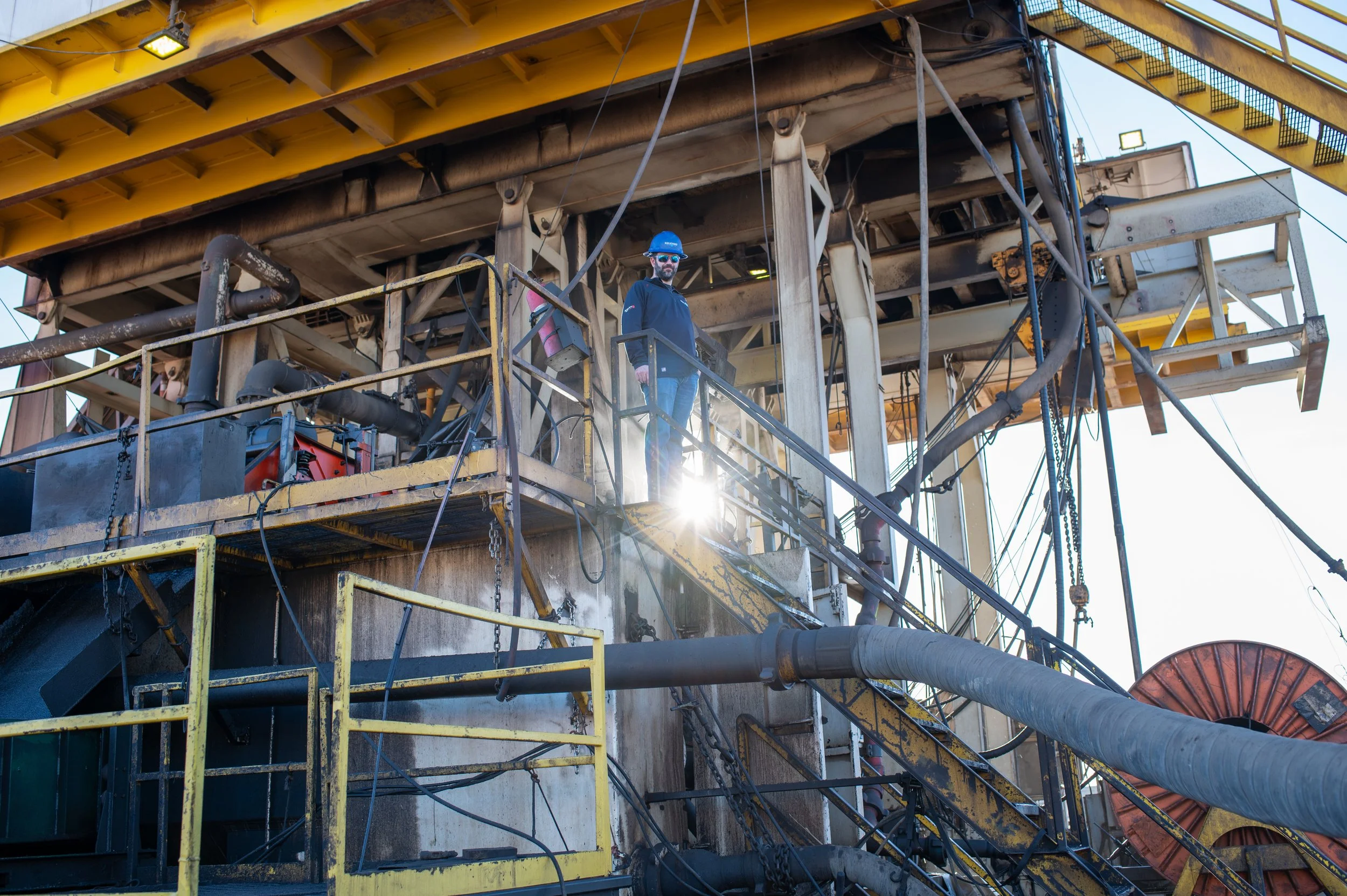 A man wearing safety gear, including a blue helmet and sunglasses, standing on an industrial platform on an oil drilling rig, with pipes, machinery, and scaffolding around him.