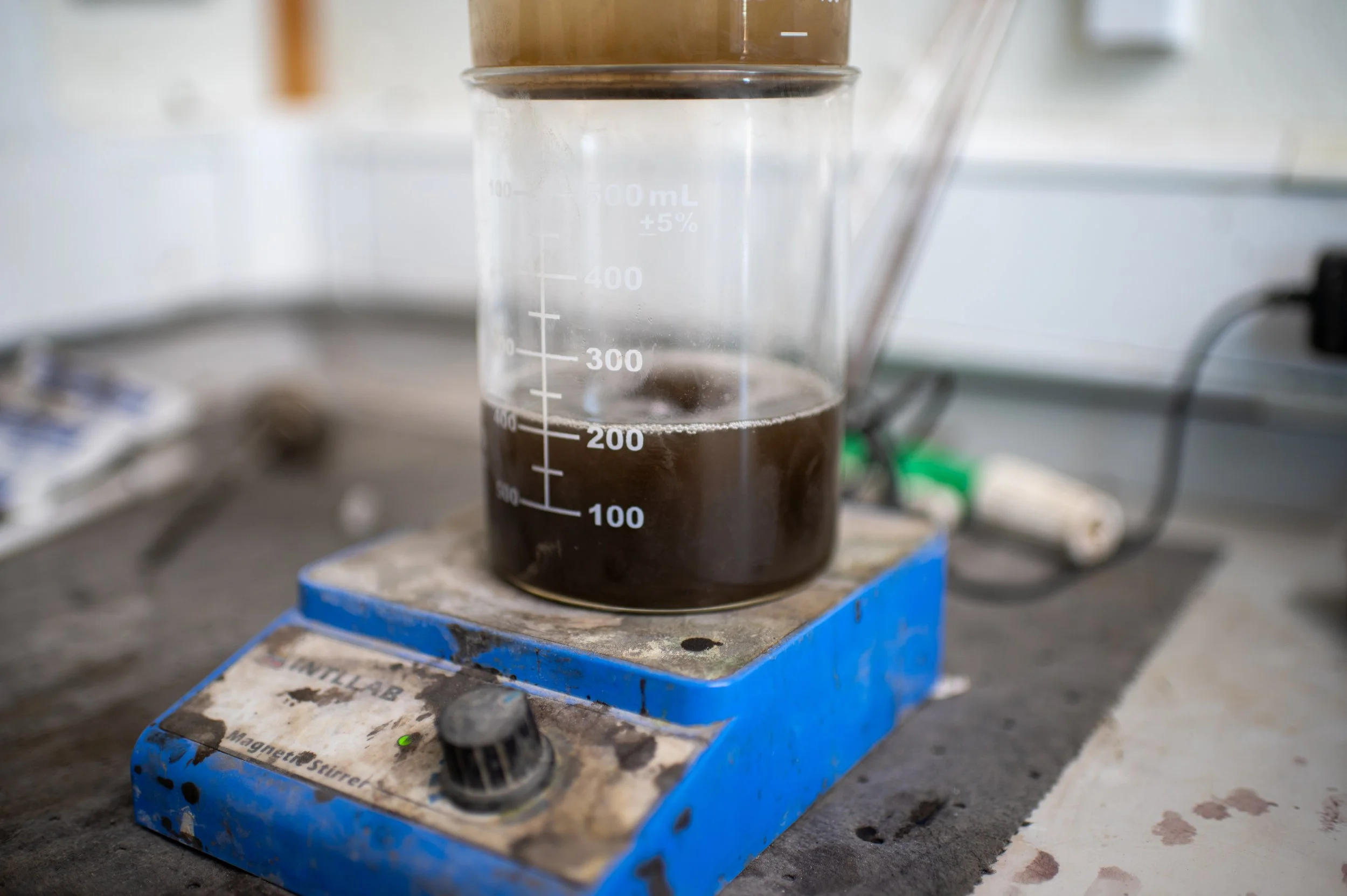 A glass beaker with dark liquid on a magnetic stirrer in a laboratory setting.