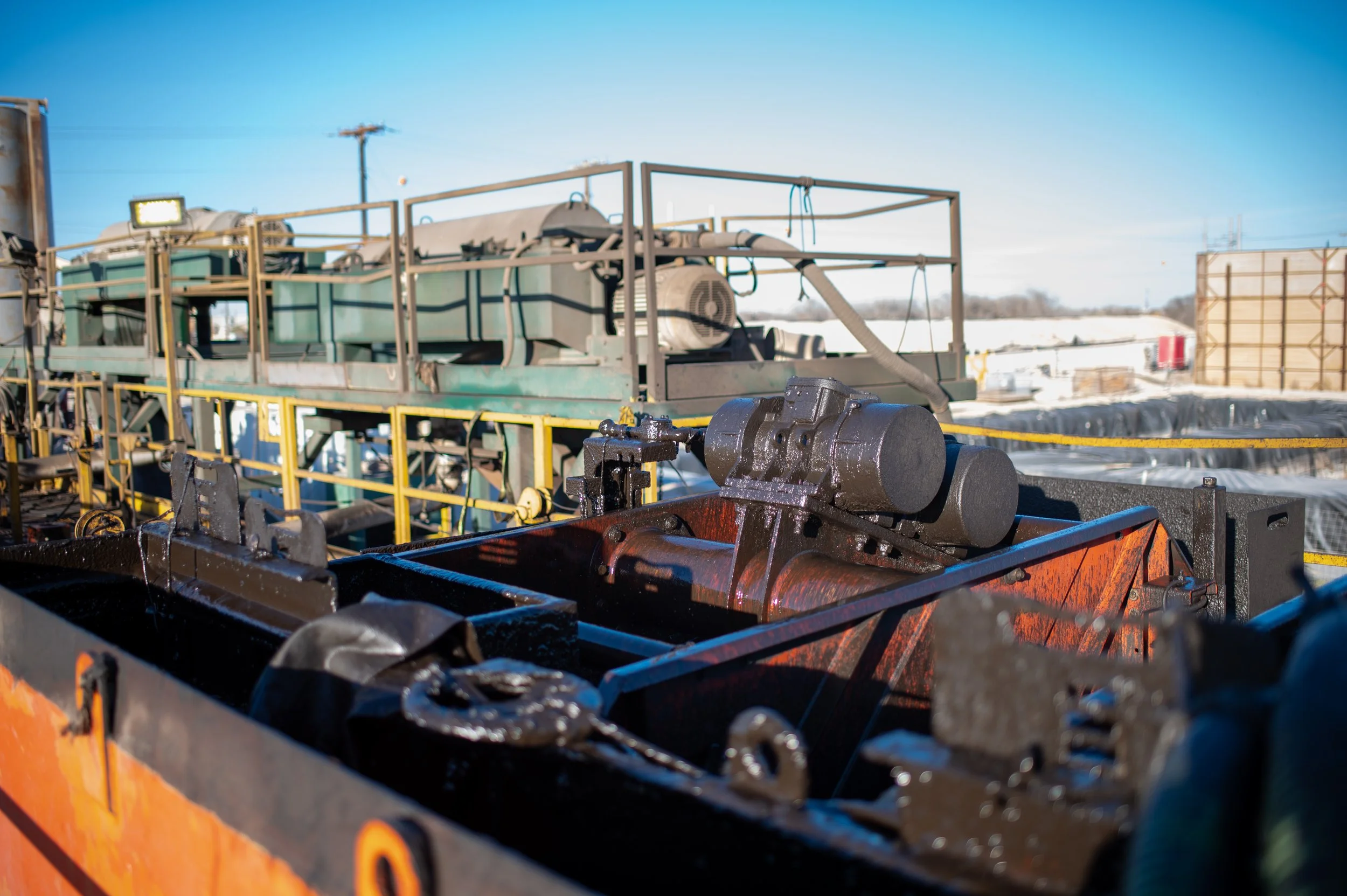Industrial machinery with pipes, tanks, and railings outdoors on a sunny day with snow in the background.