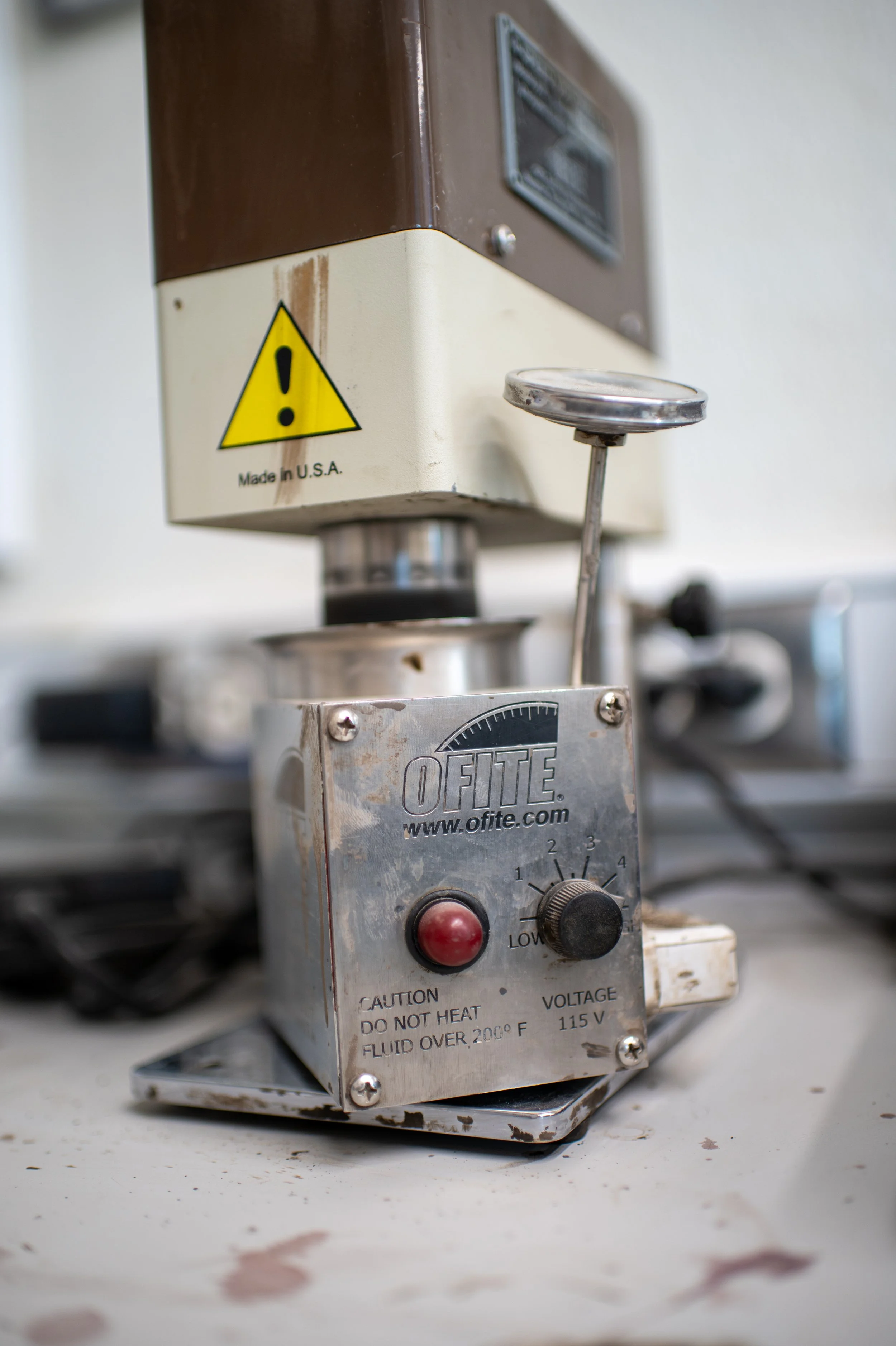 Close-up of a laboratory or industrial machine with a metal control box labeled 'OFITE' featuring a red button, a dial, and a warning sign, with a larger machine in the background.
