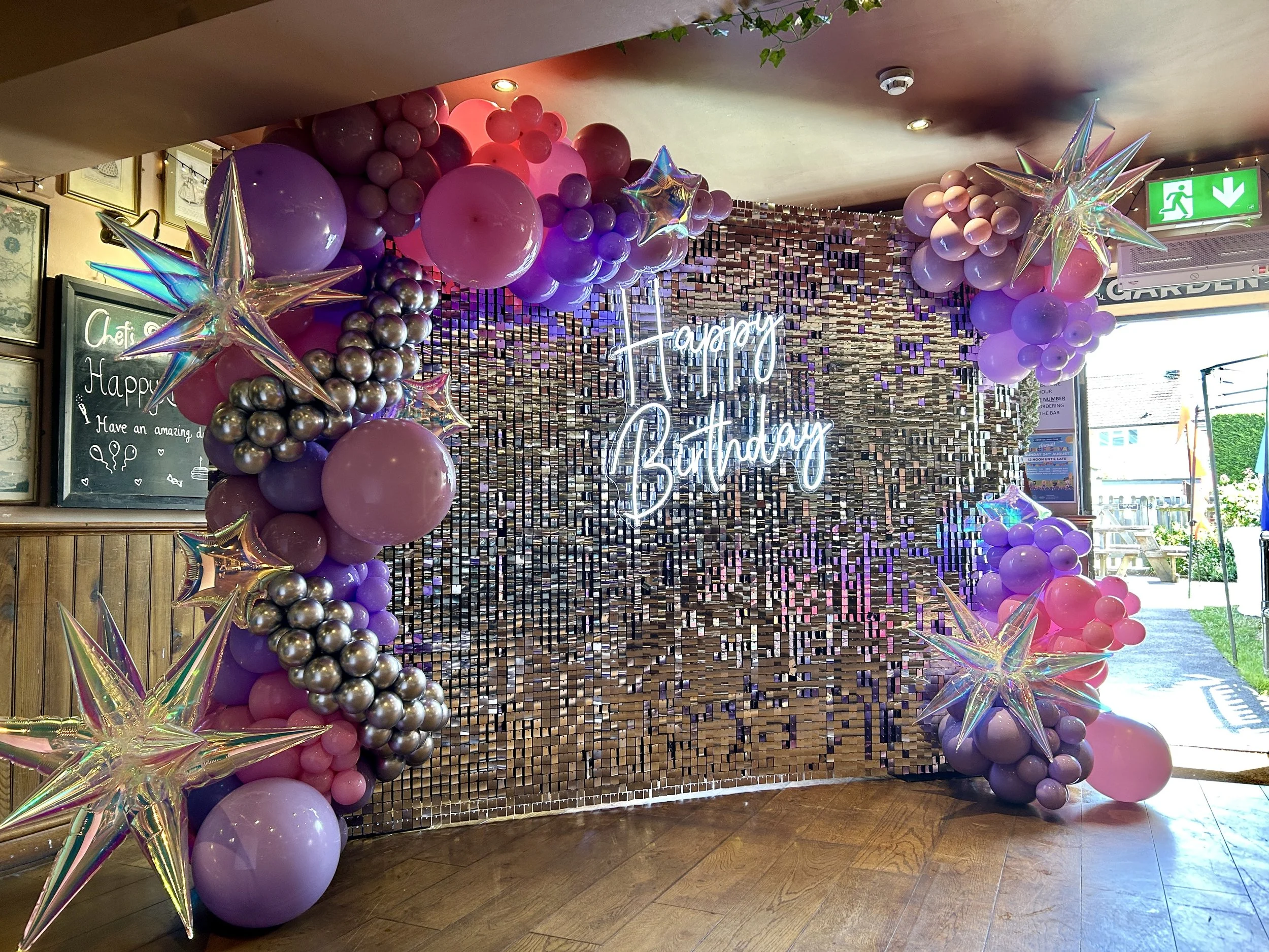 silver shimmer wall with light up happy birthday sogn in middle with a ballon garland made up of pink, lilac and silver balloons at the old runnings mill pub in east grinstead
