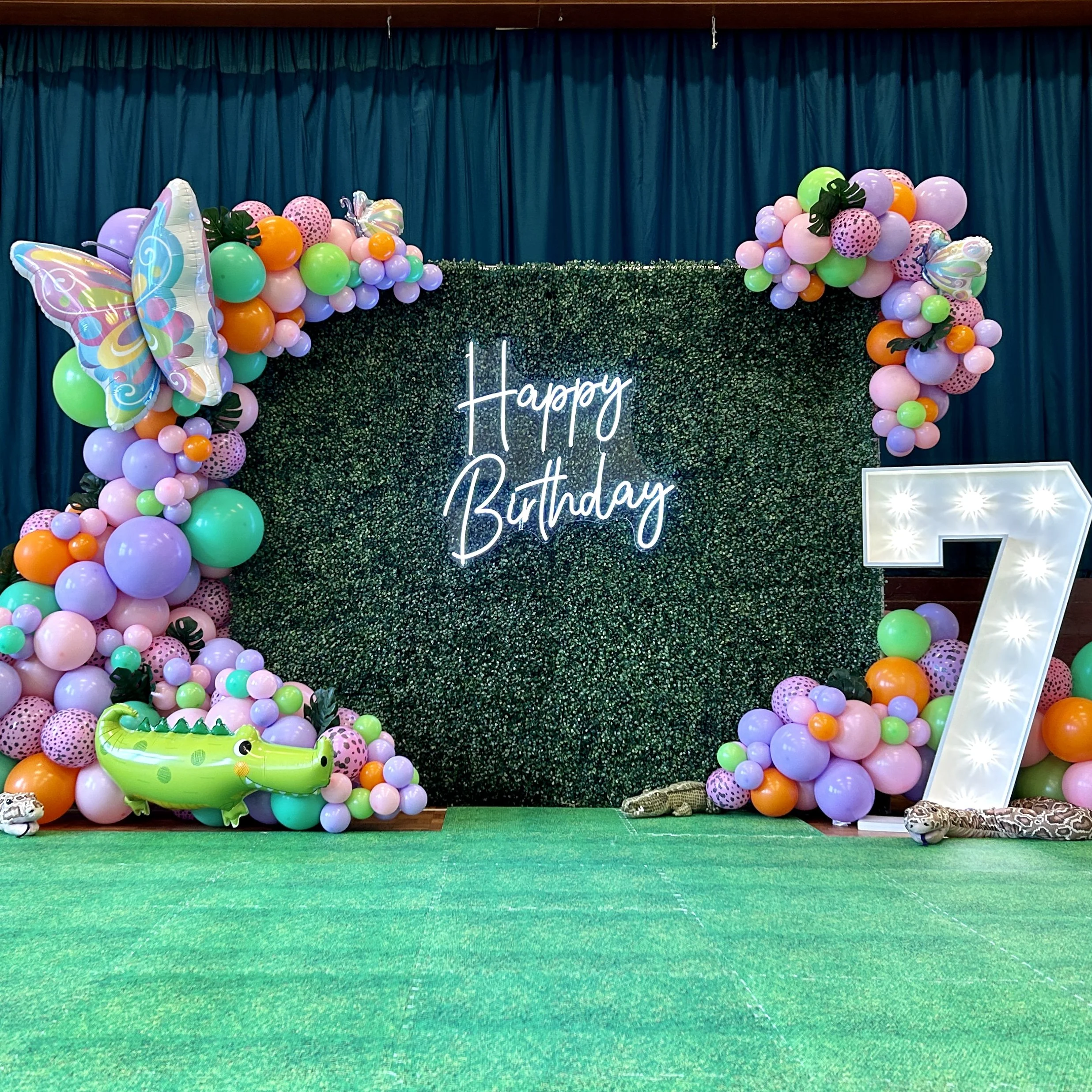 foliage wall with pink, green, orange and purple balloons a light up number 7 animal soft toys and a happy birthday sign hanging in the middle in arkley for a 7th birthday