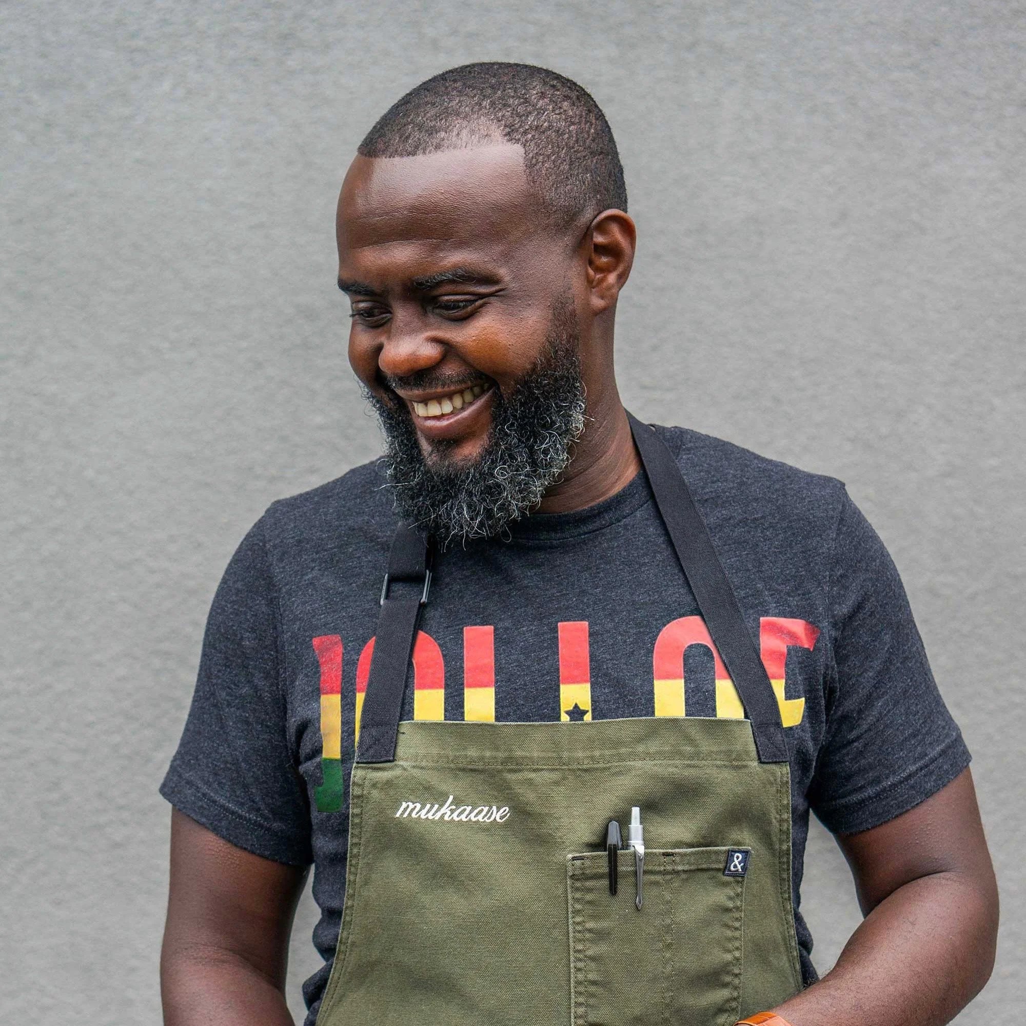 A smiling man with a short beard, wearing a black t-shirt and a gray apron, stands against a plain gray wall.