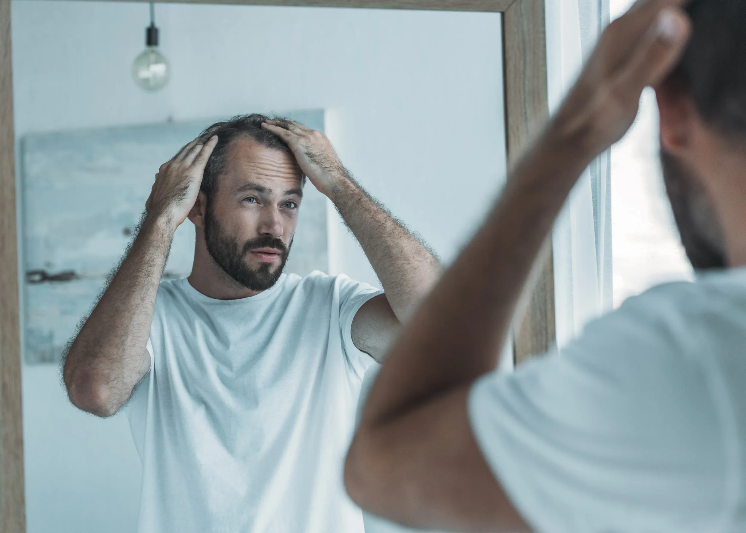A man looking at his reflection in a mirror, touching his hair with both hands, with a slightly concerned or thoughtful expression.