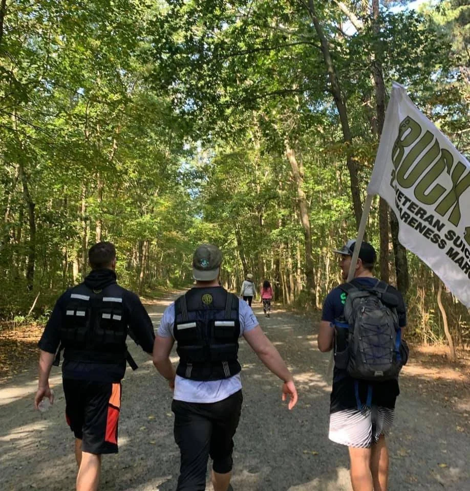 Three people walking on a forest trail. Two men in weighted vests, one carrying a "Ruck Veteran Suicide Awareness March" flag. Trees line the path, and another group is visible in the distance.