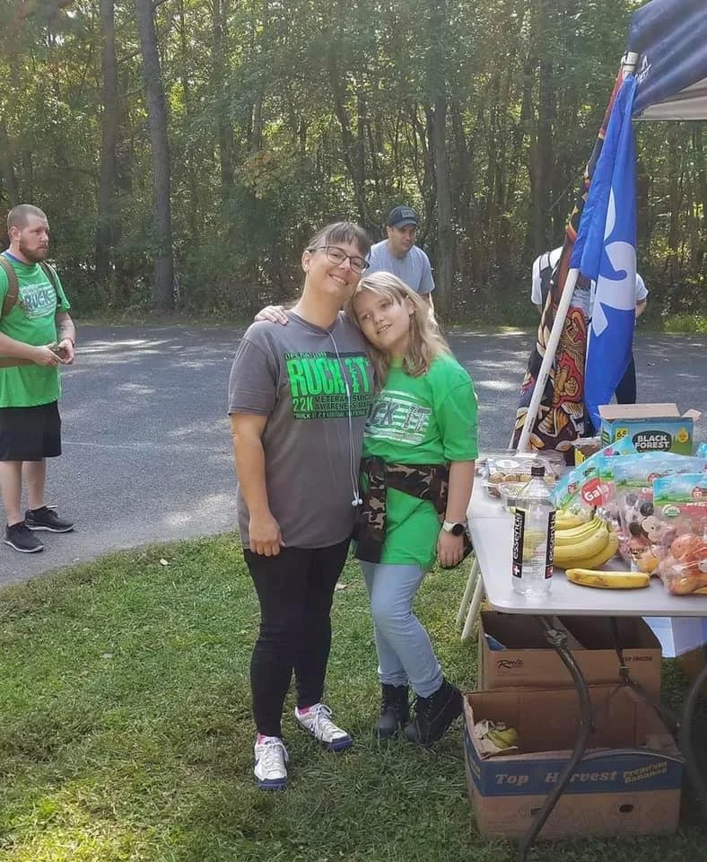 Two people standing together at an outdoor event with food items on a table beside them. They are both wearing matching event shirts in green and gray. Other participants can be seen in the background, and a flag is visible near the table.
