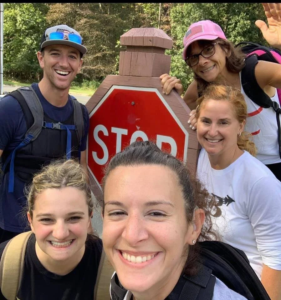 Group of five people smiling and posing next to a wooden stop sign in an outdoor setting with trees in the background.