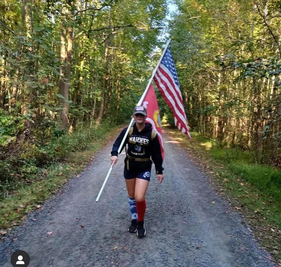 Person walking on a wooded trail, wearing a "Marines" sweatshirt, holding a large American flag and another flag with a red design. Wearing shorts and mismatched socks. Trees line both sides of the path.