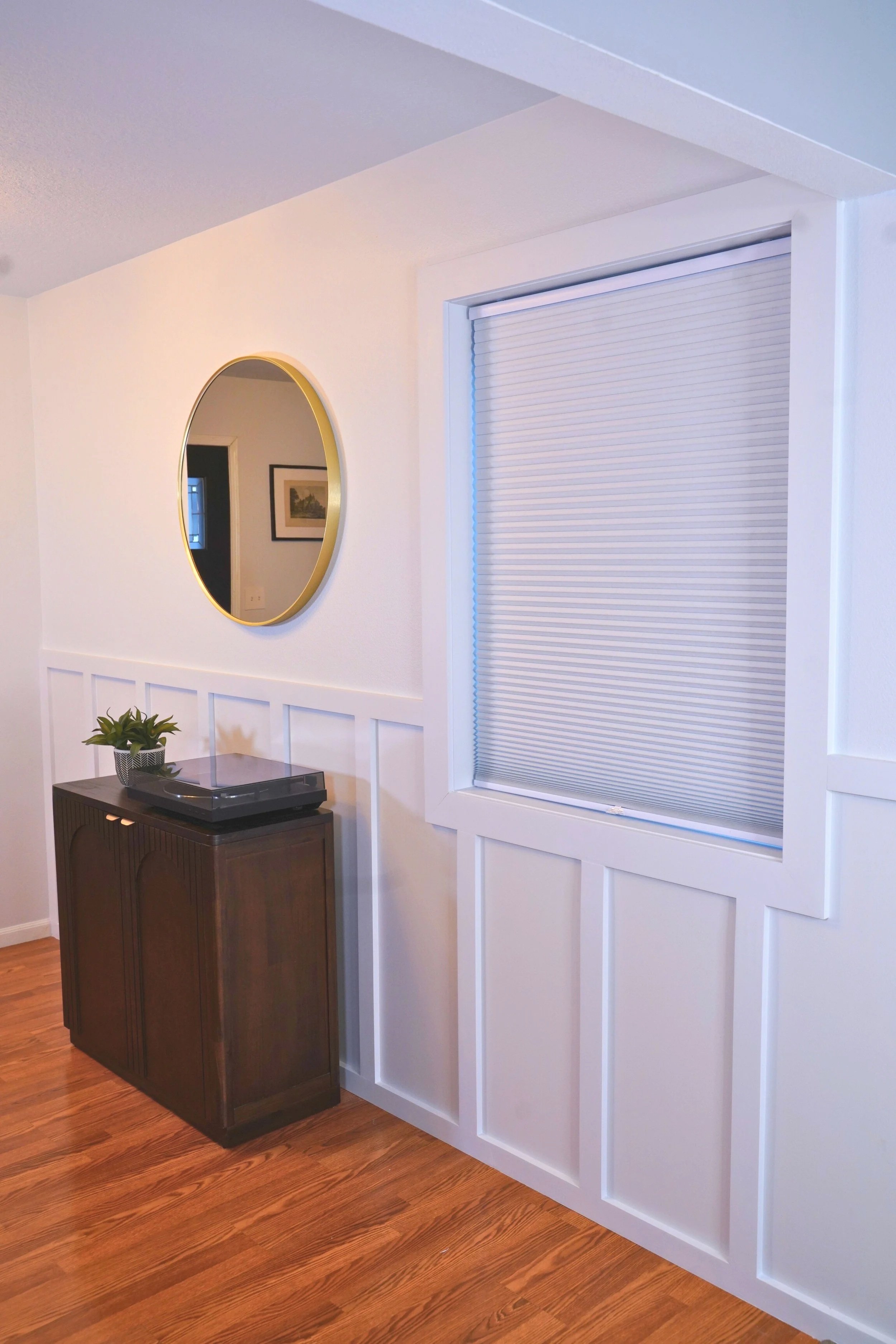 Interior of a room with wood flooring, a white wall with decorative paneling, a window with closed blinds, a dark wooden cabinet with a record player and potted plant on it, a round mirror on the wall reflecting a framed picture and a doorway.