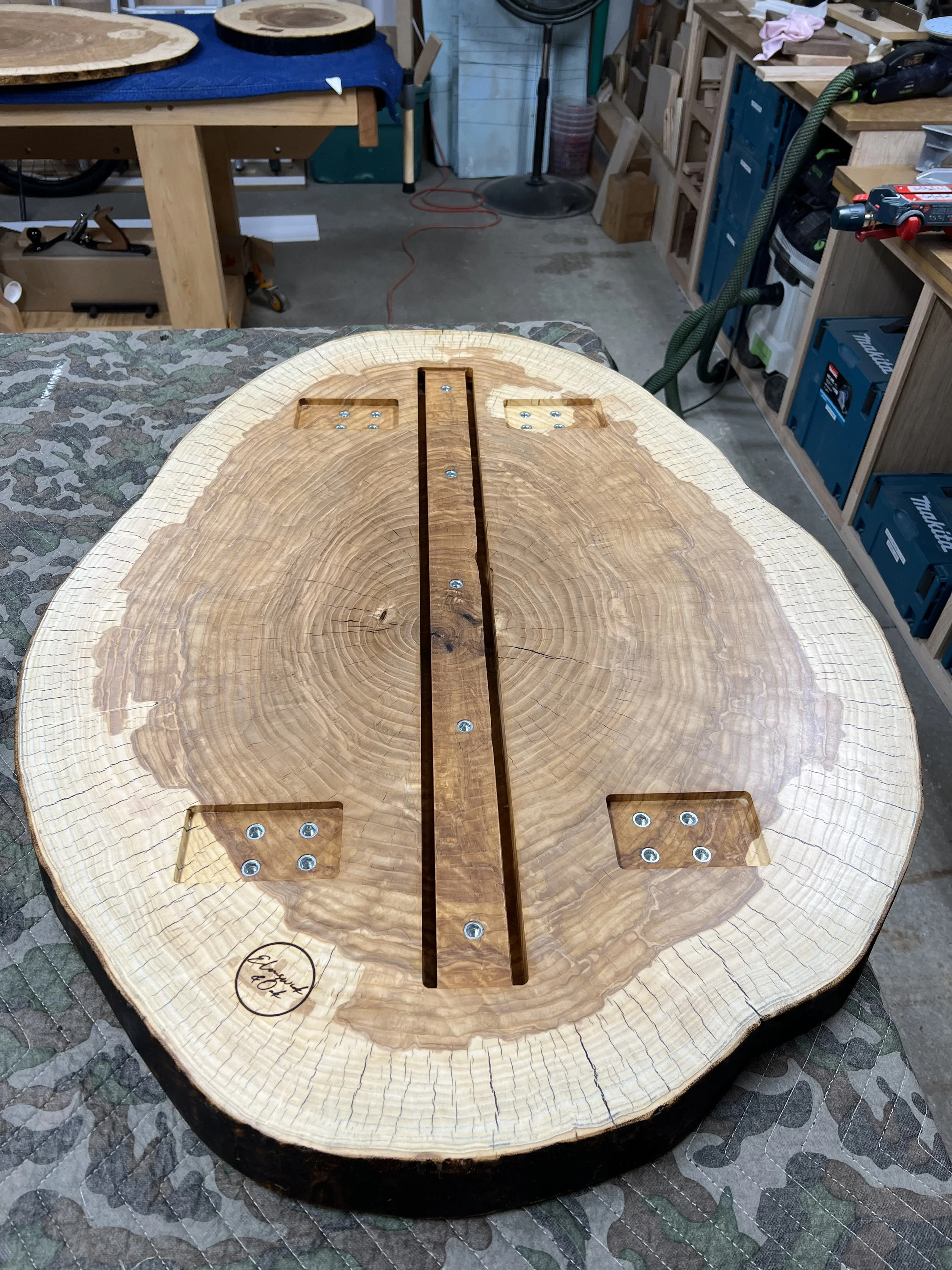 A wooden coffee table with a natural edge, displaying a decorative inlay of darker wood running lengthwise through the center. The tabletop has a polished finish, showcasing the natural grain and growth rings of the wood. In the background, a woodworking workshop is visible with workbenches, tools, and materials.