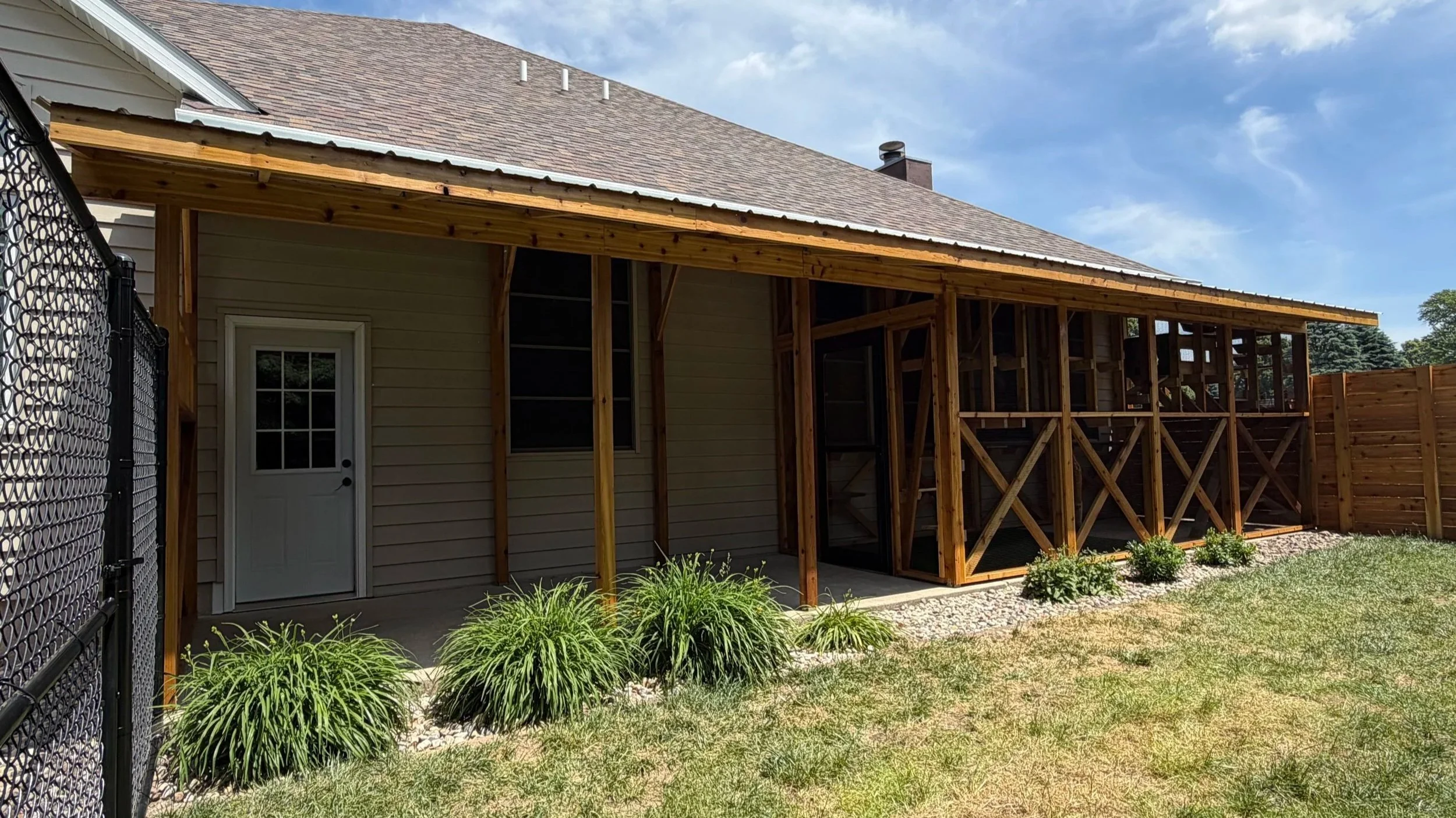 Backyard of a house with an added covered patio and catio, surrounded by a wooden fence and landscaped with green bushes and grass.