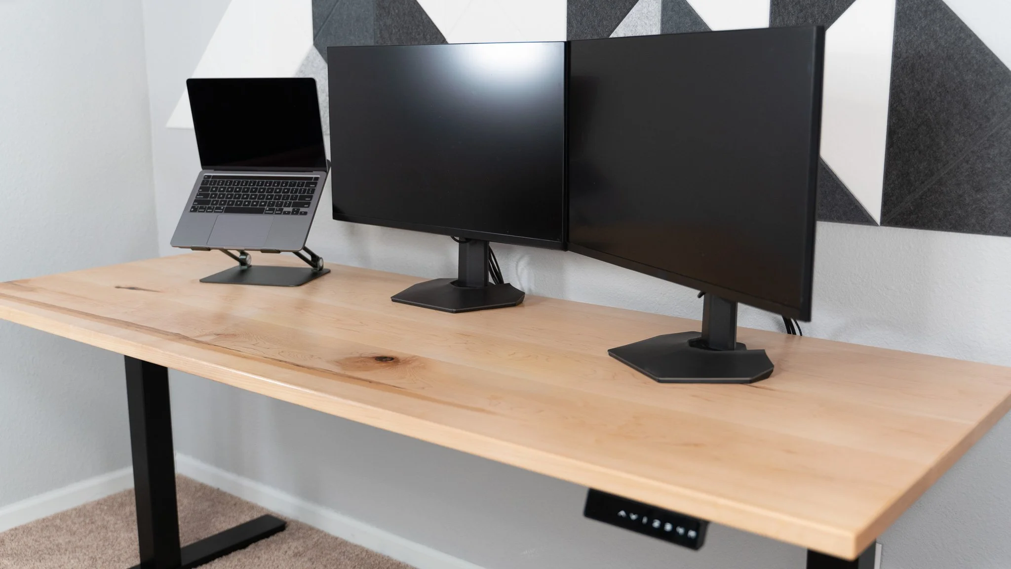 A wooden desk with three computer monitors mounted on stands and a closed laptop on a stand, set against a wall with soundproofing foam panels.