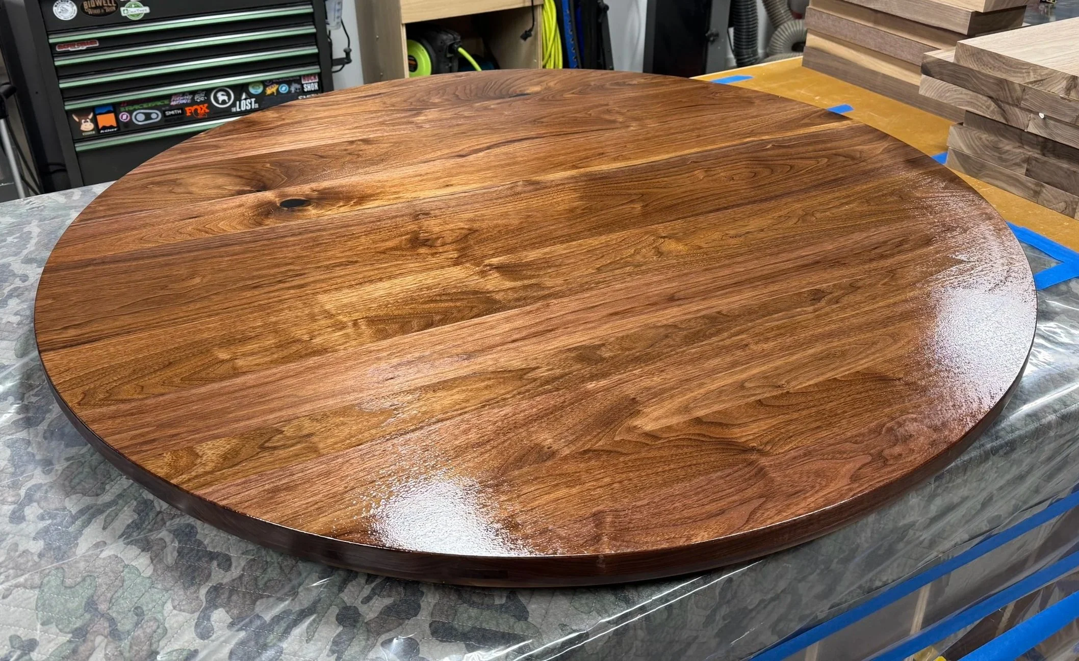 A round, polished wooden table on a workshop with tools and wood pieces in the background.