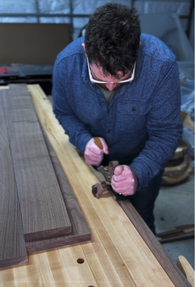 A man in glasses and a blue sweater is using a hand plane to smooth a piece of wood in a woodworking shop.