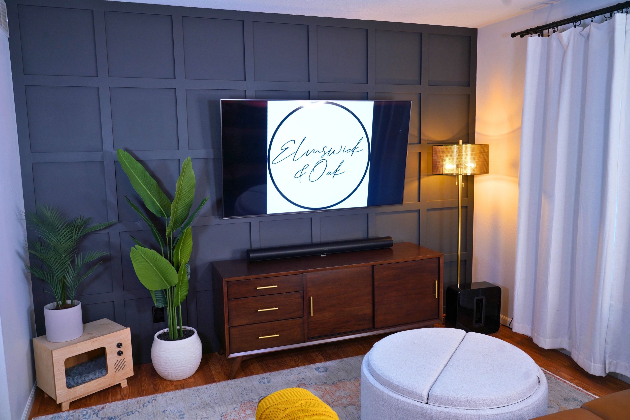 Living room with dark decorative paneling, a wall-mounted TV displaying 'Elmswick & Oak' logo, dark wood console, tall green plants in white pots, gold floor lamp, white curtains, and a round beige ottoman.