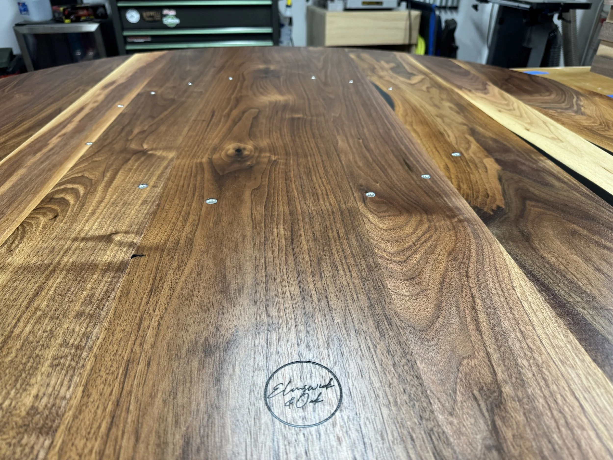 Close-up photograph of a wooden table with a natural finish, showing the grain and color variations of the wood. There is a handwritten signature in black ink with the words 'Elizabeth Pox' within a circle at the bottom of the image.