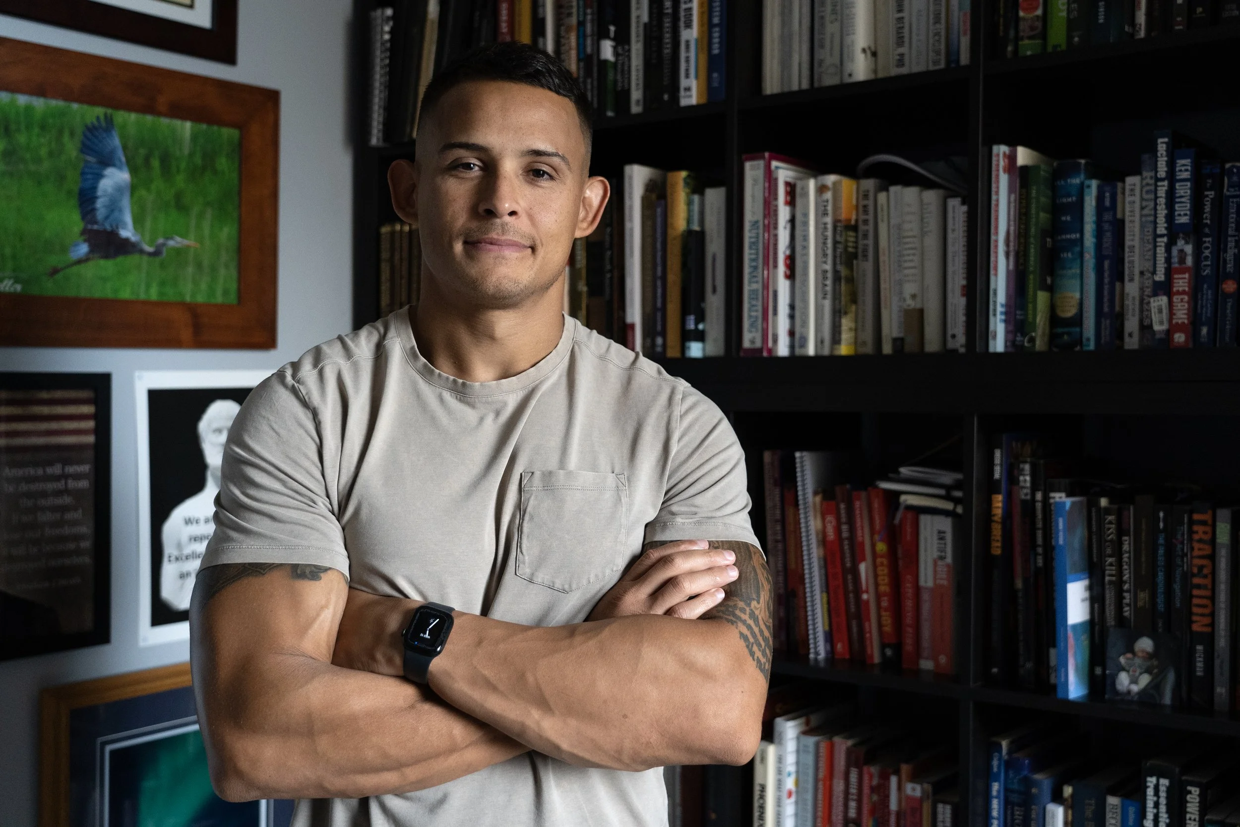 A man with short dark hair, wearing a beige t-shirt and a black smartwatch, standing with arms crossed in front of a bookshelf filled with books, in a room with framed pictures and a colorful bird poster on the wall.