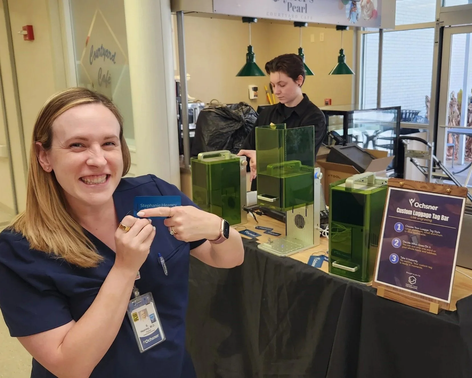 A woman with shoulder-length blonde hair, wearing a navy blue uniform and a name badge, smiling and pointing at her luggag tag. She is at a luggage tag bar station with green equipment and an informational sign, in a brightly lit indoor setting.