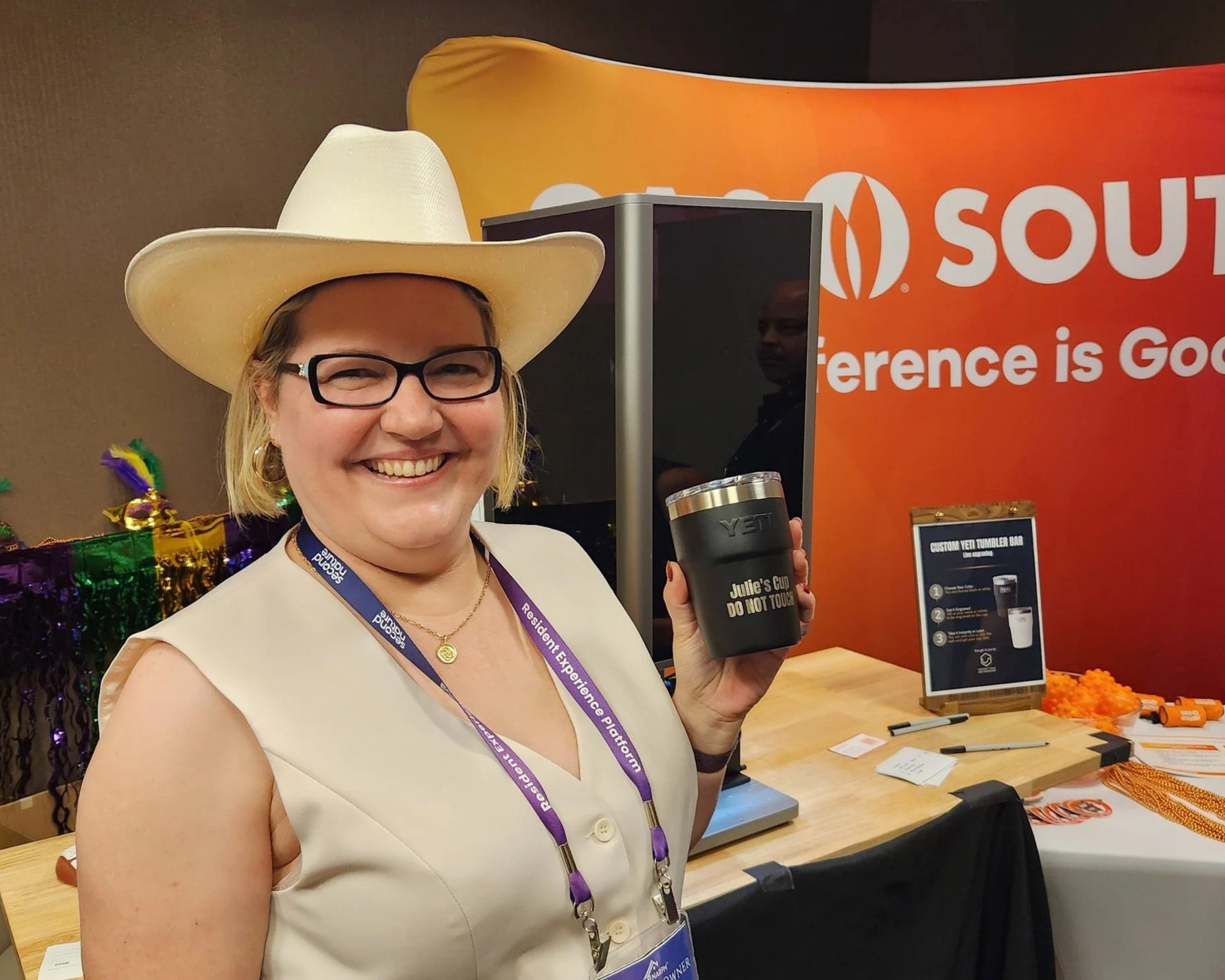 A woman wearing a white sleeveless blazer, black glasses, a cowboy hat, and a purple lanyard smiling at the camera, holding a tumbler that reads "Julie’s Cup, Do Not Touch," in an indoor setting with a colorful background and promotional materials on a table.