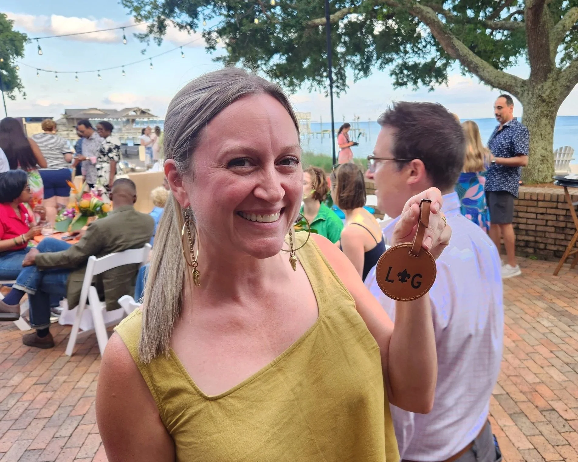 Close-up of a smiling woman with long blonde hair, wearing a yellow top and earrings, holding a leather item with 'L G' embossed on it. In the background, an outdoor gathering near a body of water with people, trees, and string lights.