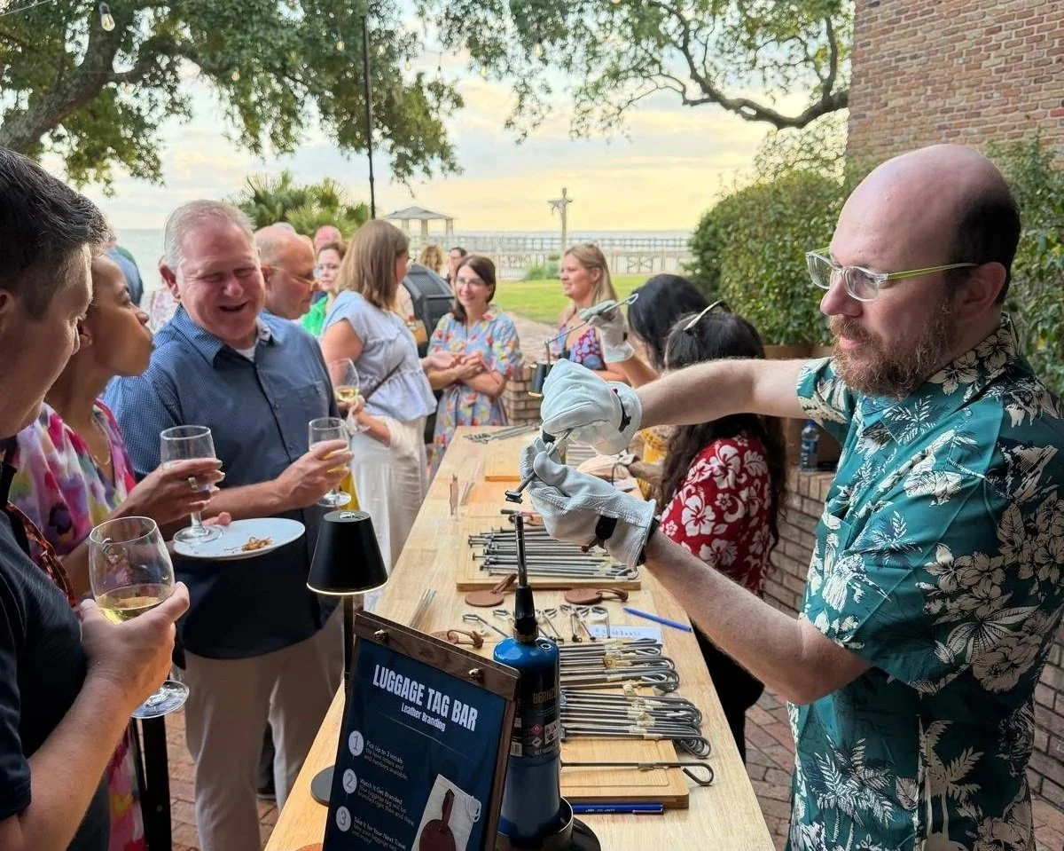 People attending an outdoor event with a luggage tag bar, some holding drinks, as a man in a Hawaiian shirt prepares items behind a bar table with tools and a sign.