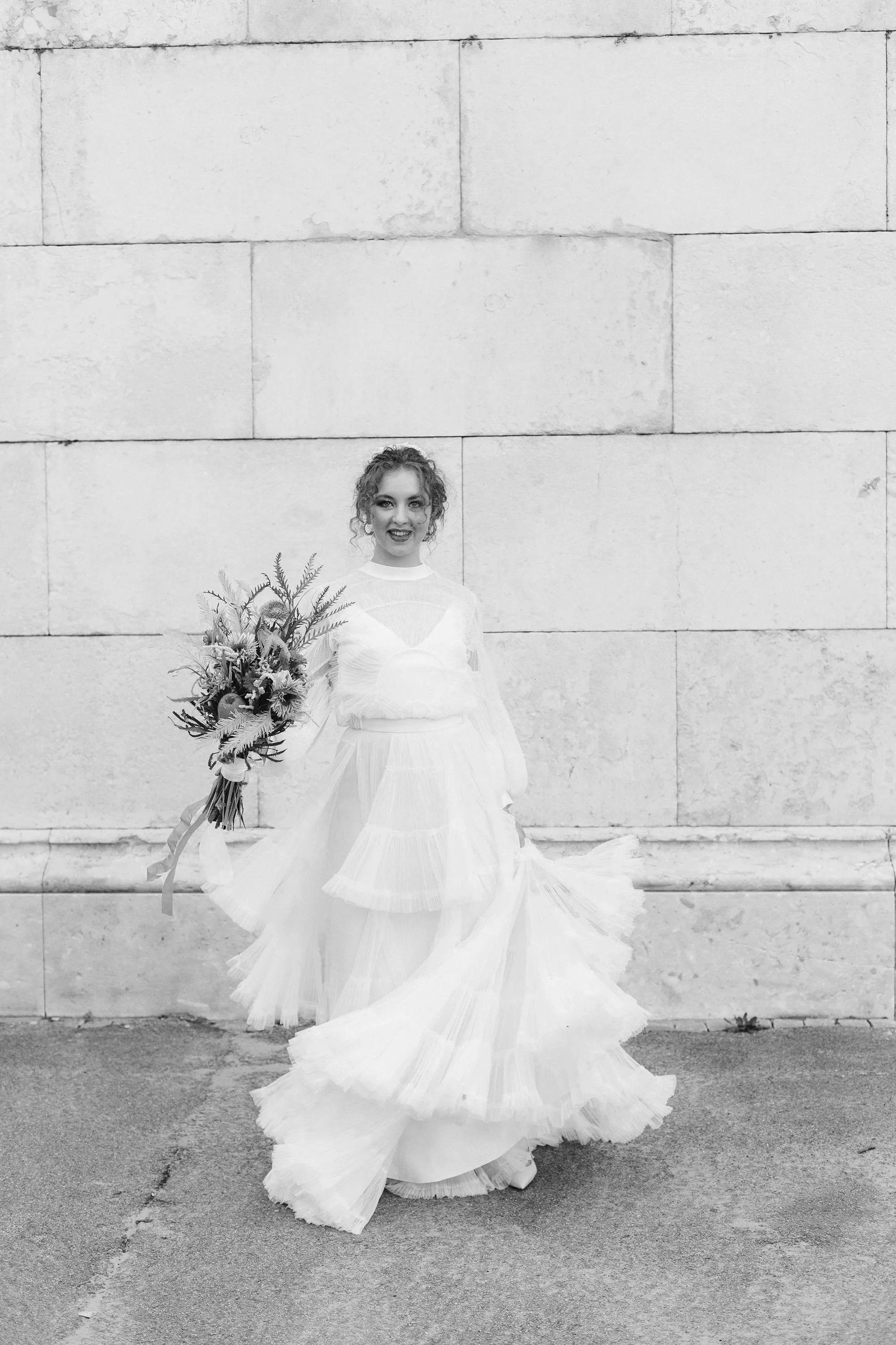 A woman in a white wedding dress holding a bouquet, standing against a large stone wall.