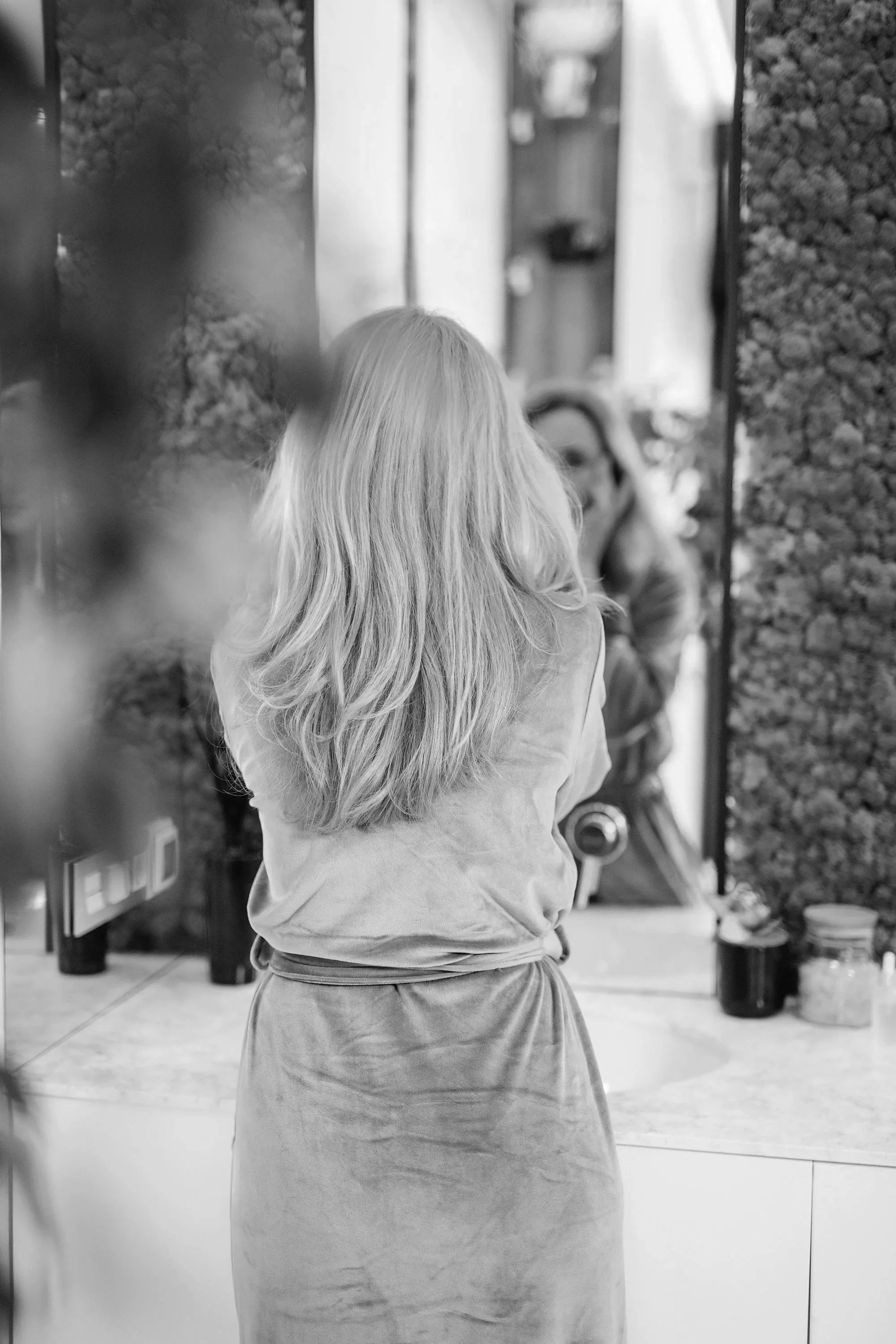 Woman with blond hair wearing a pink robe standing in front of a bathroom mirror with green plant decor.