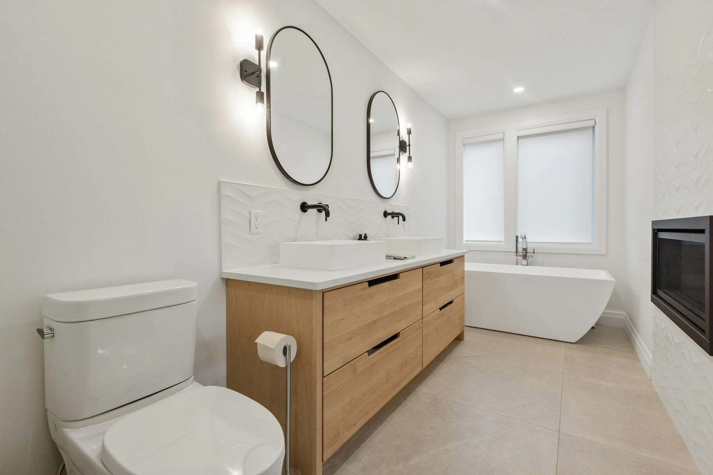 Modern bathroom with white walls, two oval mirrors, black wall-mounted light fixtures, two white vessel sinks on a wooden vanity, a toilet, a bathtub, and a microwave, with large windows letting in natural light.