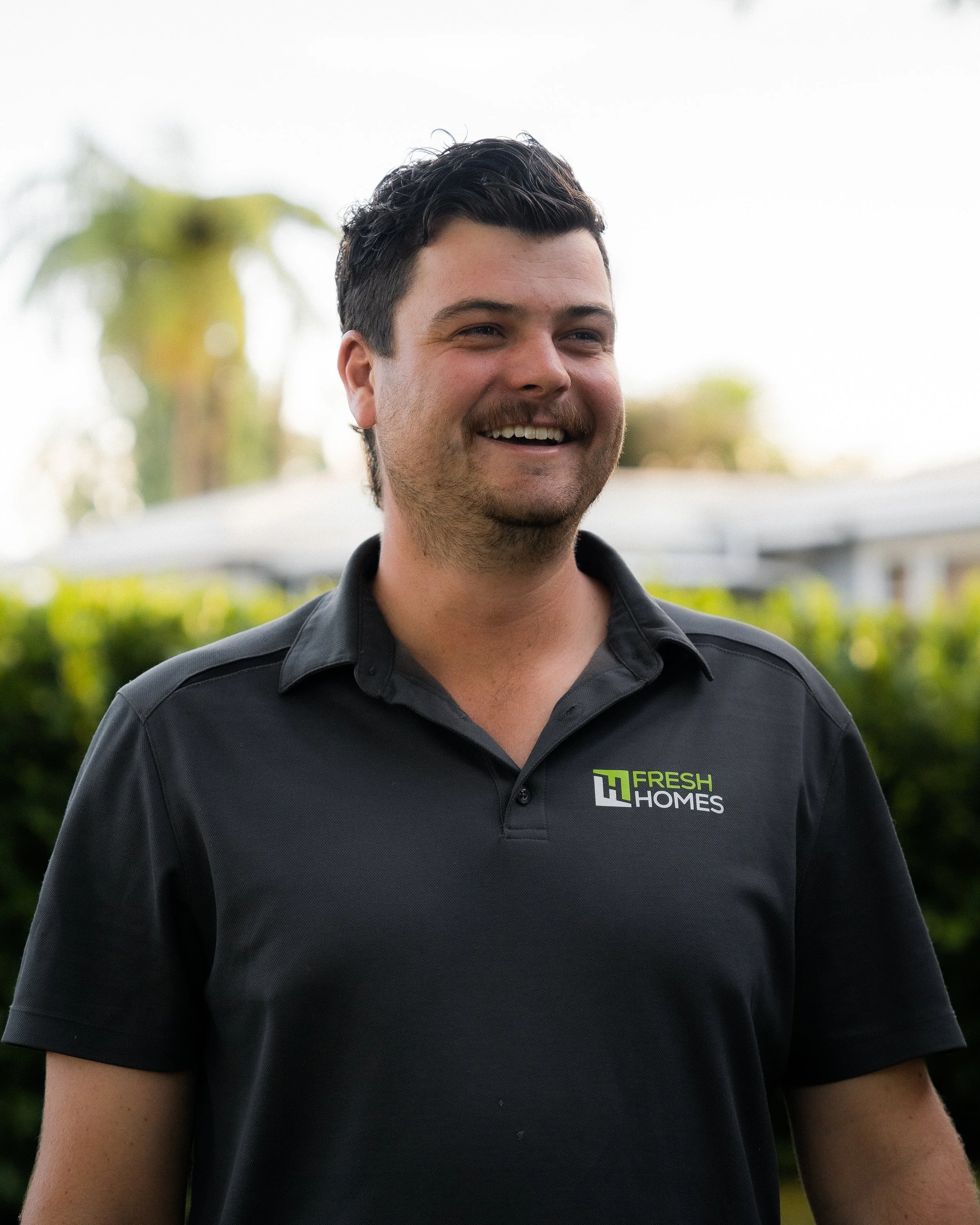 A smiling man wearing a black polo shirt with 'FRESH HOMES' logo, standing outdoors with blurred greenery and trees in the background.